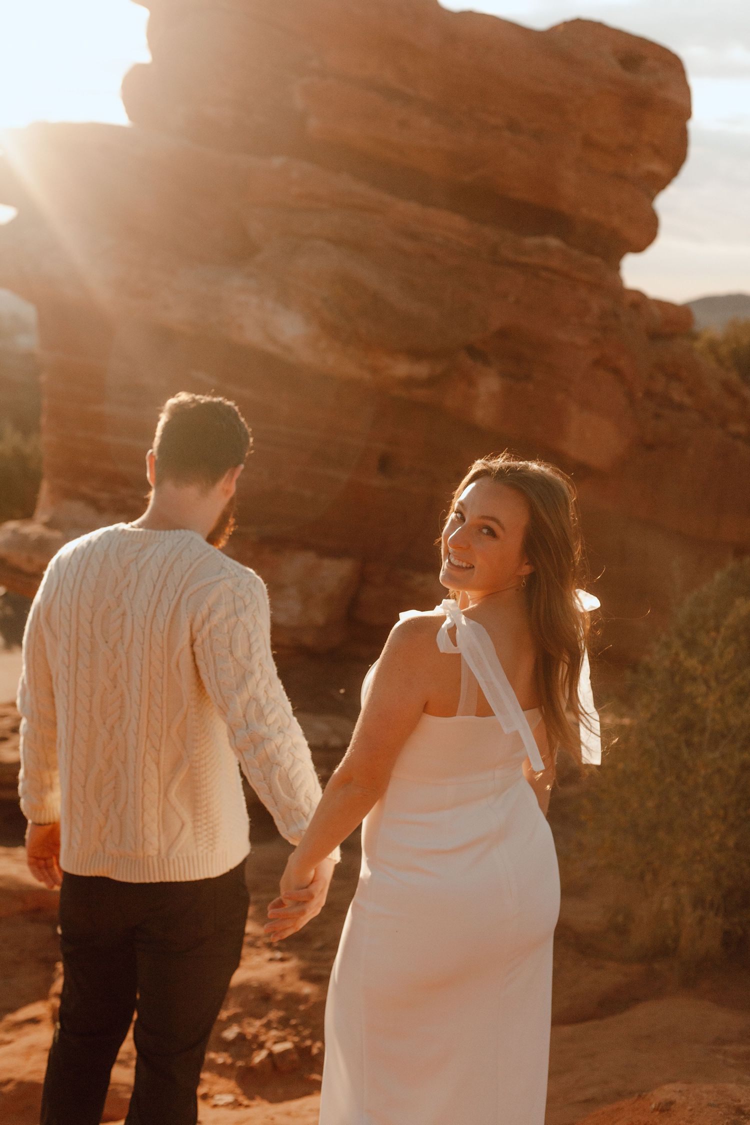 Couple holding hands walks through red rock formations at sunset.