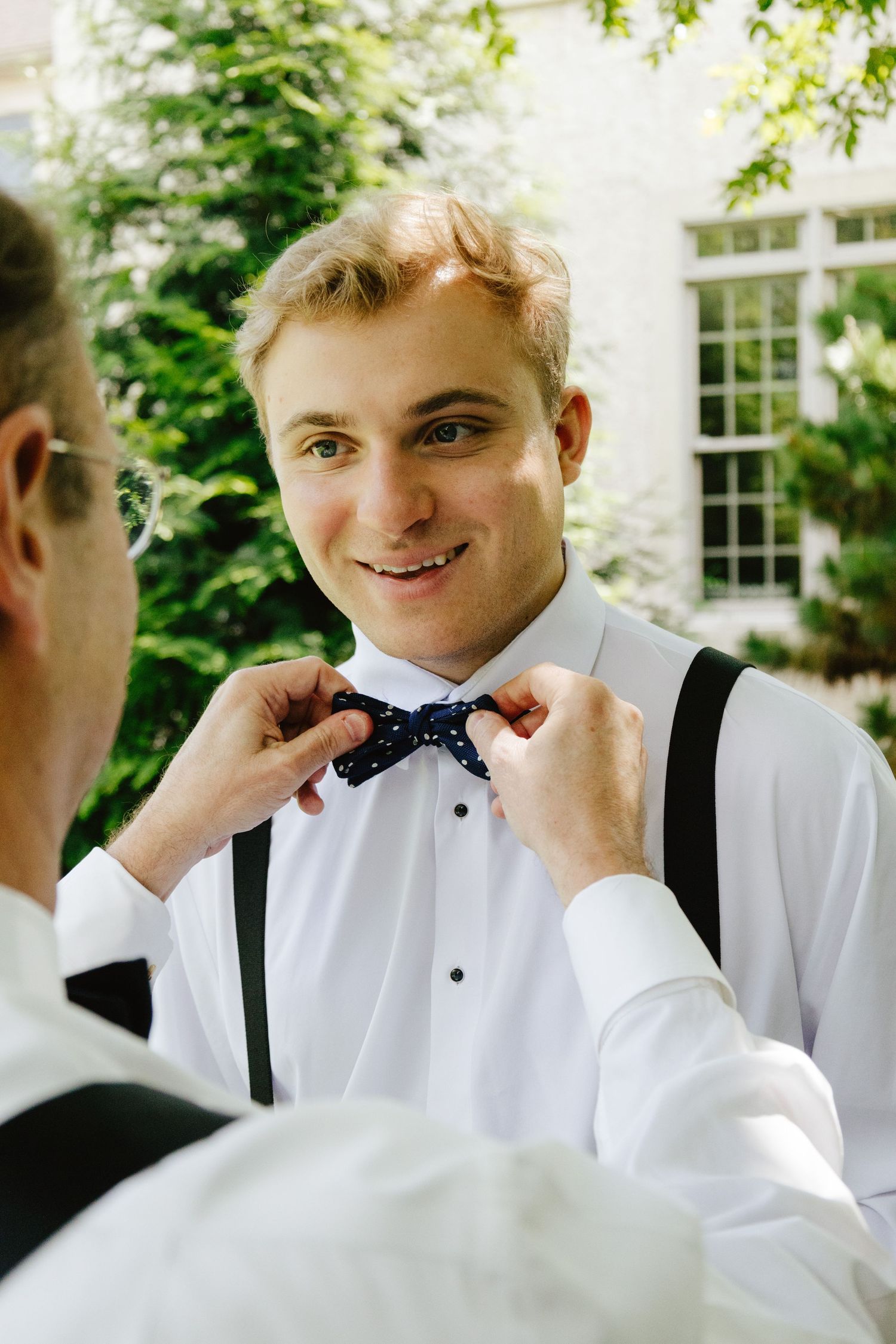 Man in formalwear with suspenders adjusts his bowtie in a series of outdoor photos with greenery in background.