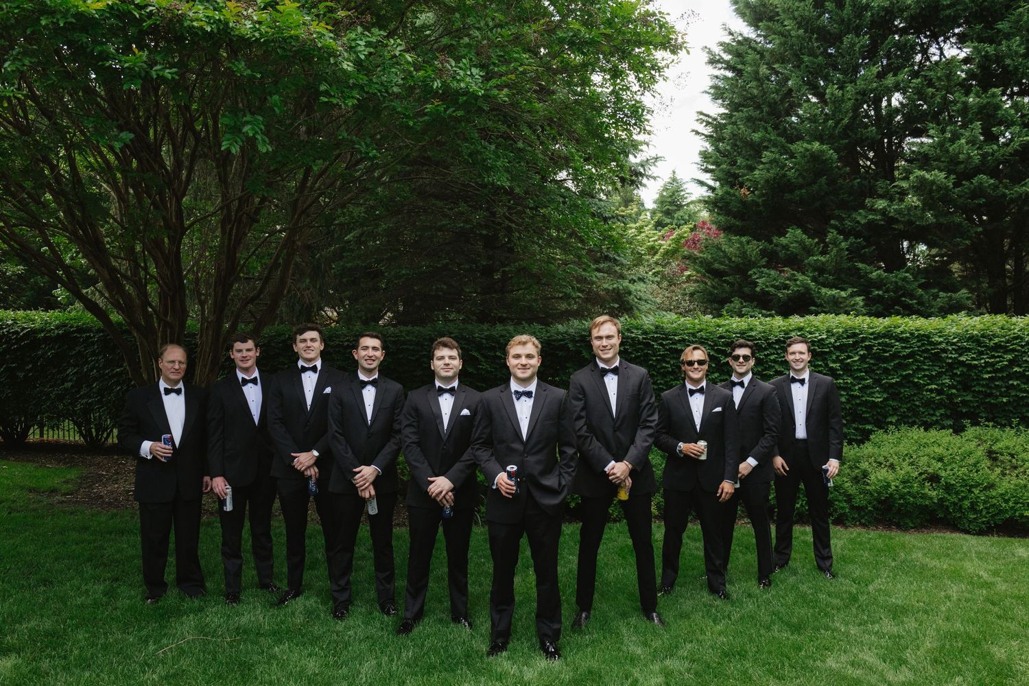 Groomsmen in matching black tuxedos pose in formation on a green lawn with landscaped hedges.