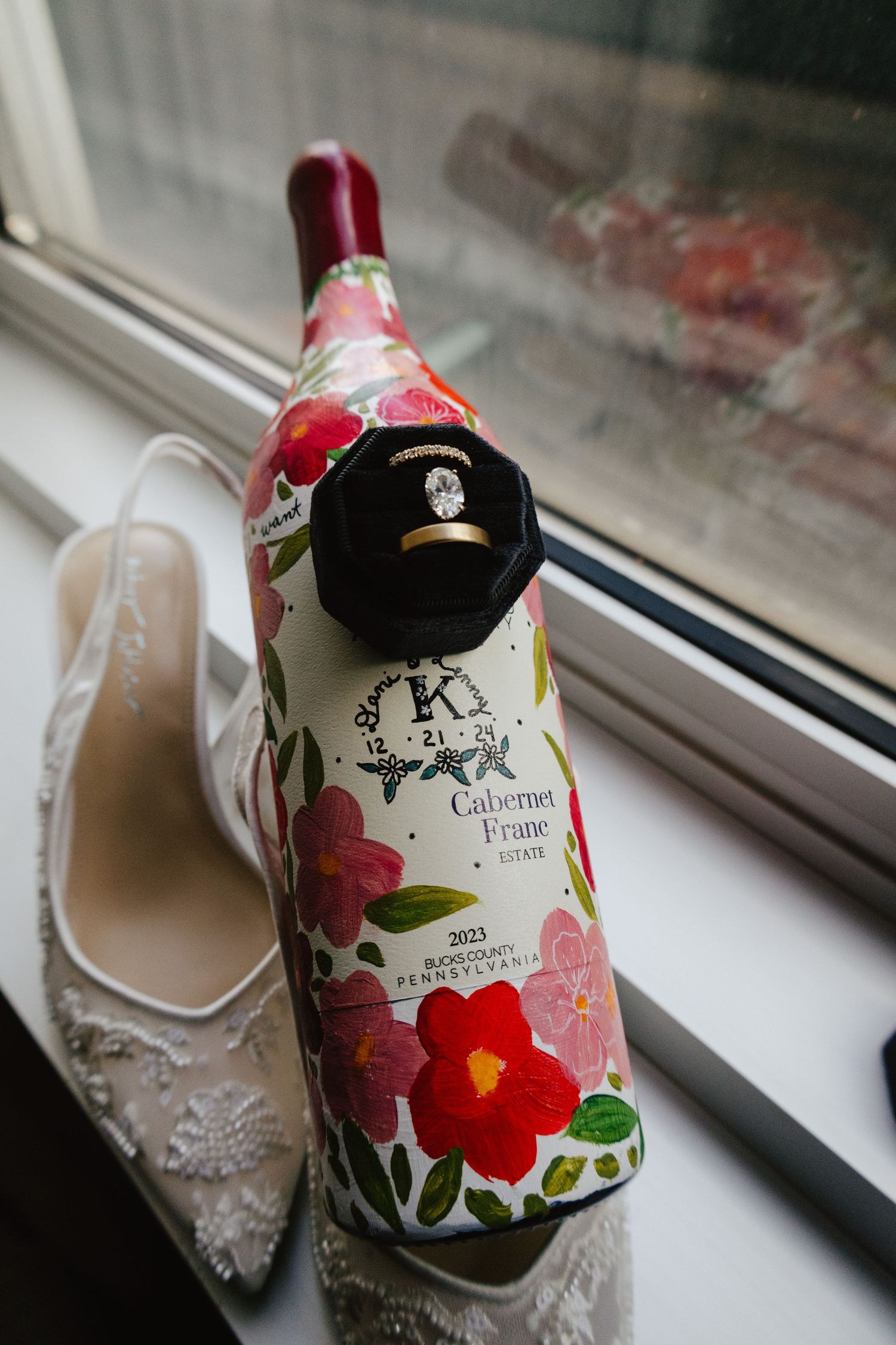 Floral decorated wine bottle next to lace wedding shoes on windowsill.