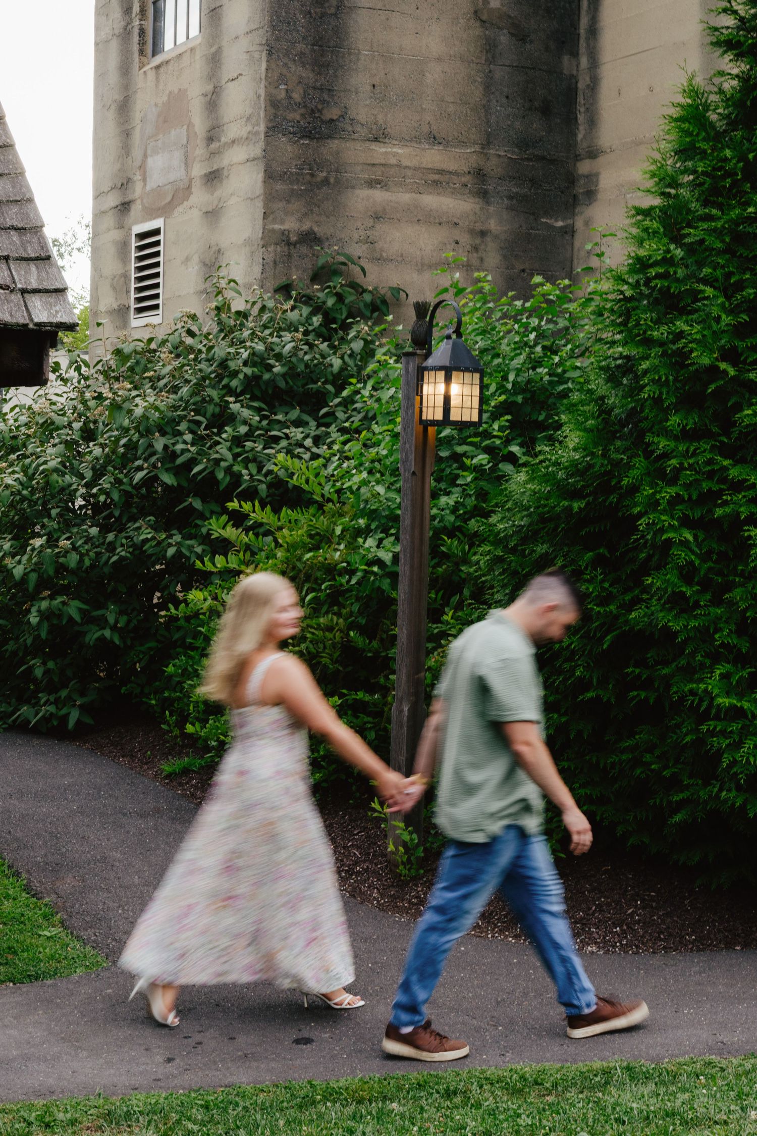 Motion blur sequence of two people walking past greenery and lamp posts on a stone pathway at dusk.