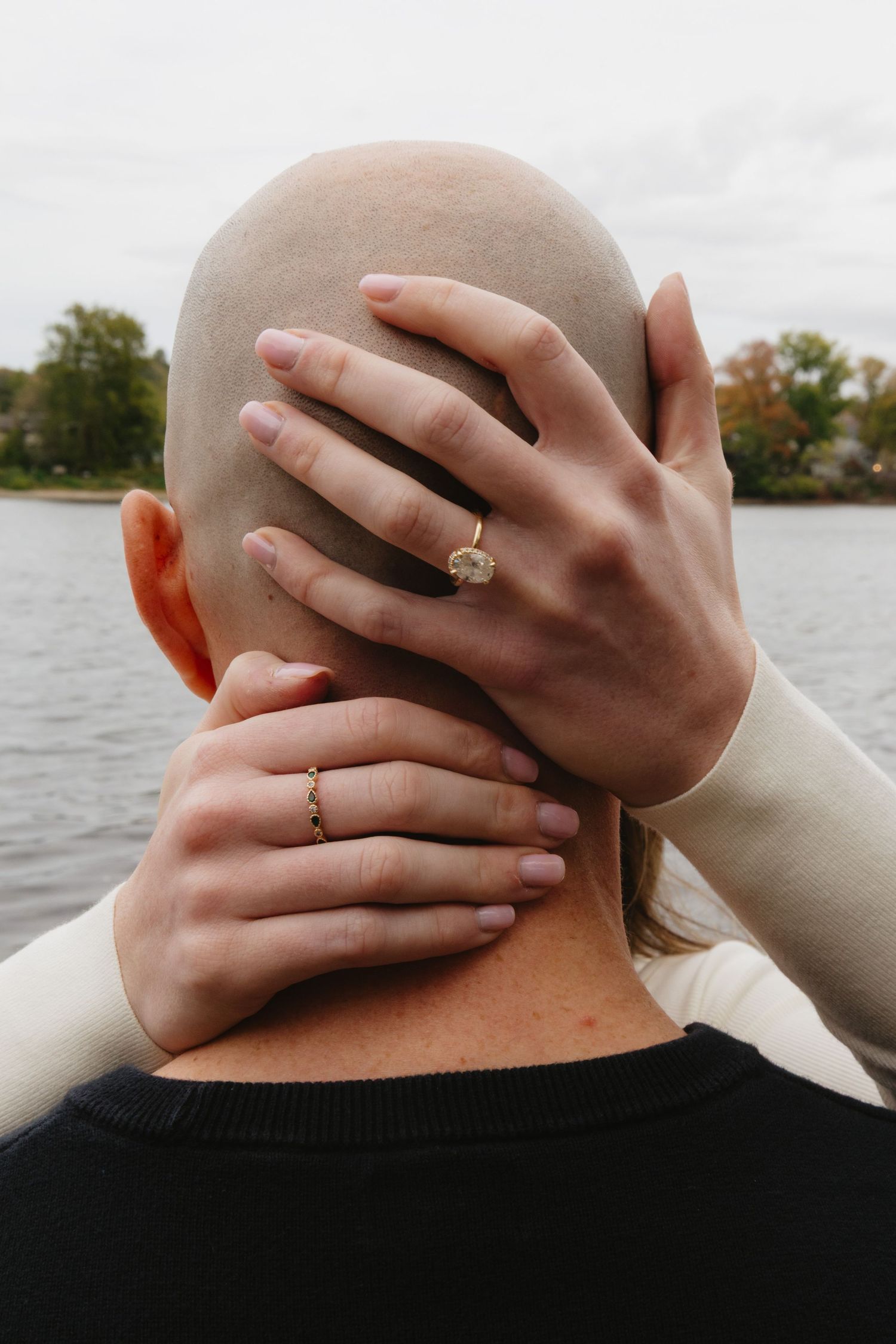 Hands with engagement rings embrace the back of a head against a waterfront background.