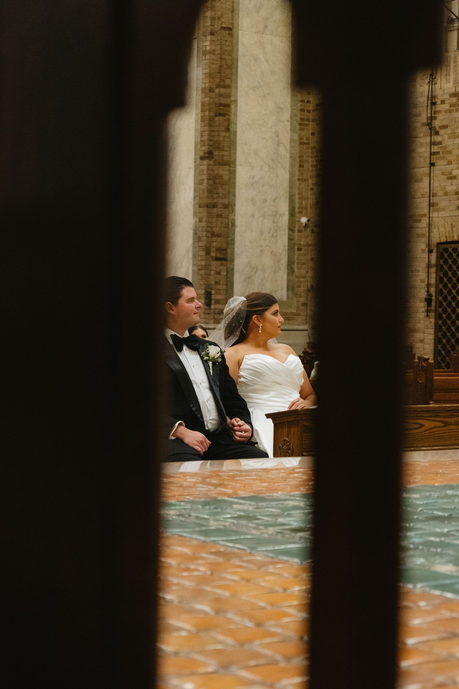 A bride and groom stand together in formal attire viewed through architectural columns at their wedding venue.