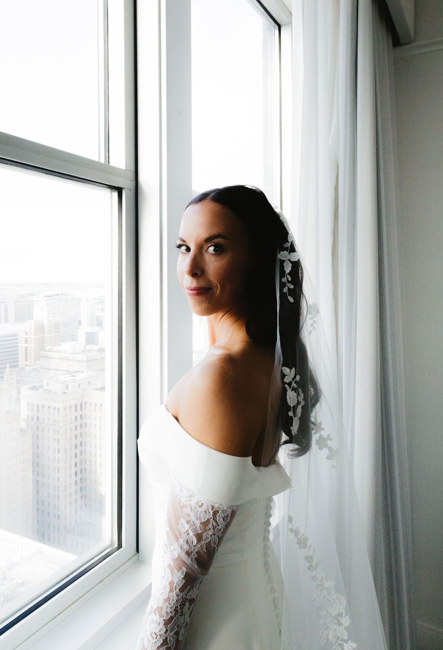 An elegant bride in a white lace gown gazes through a high-rise window.