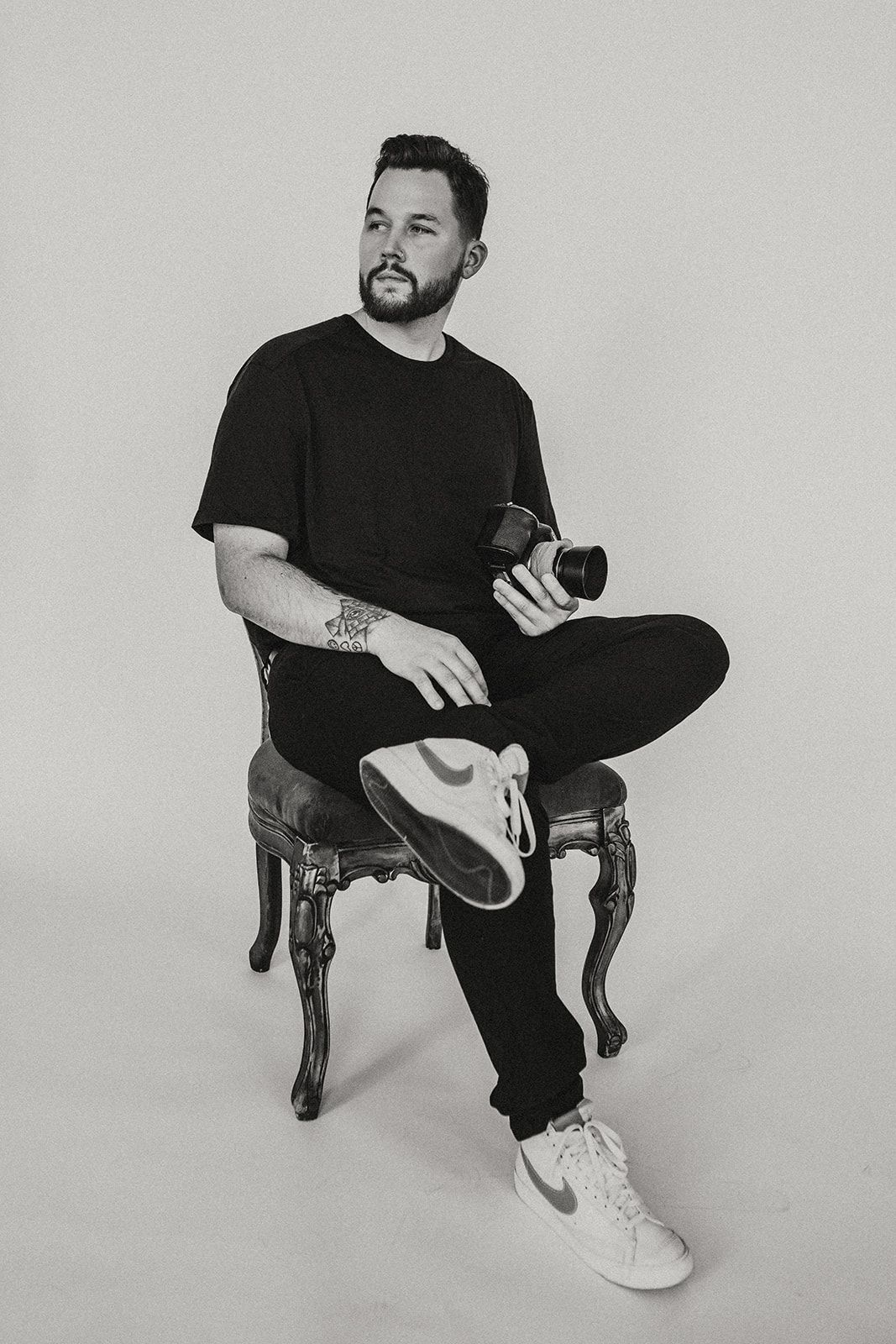 Portrait of a photographer in black attire sitting on an ornate chair holding a camera.
