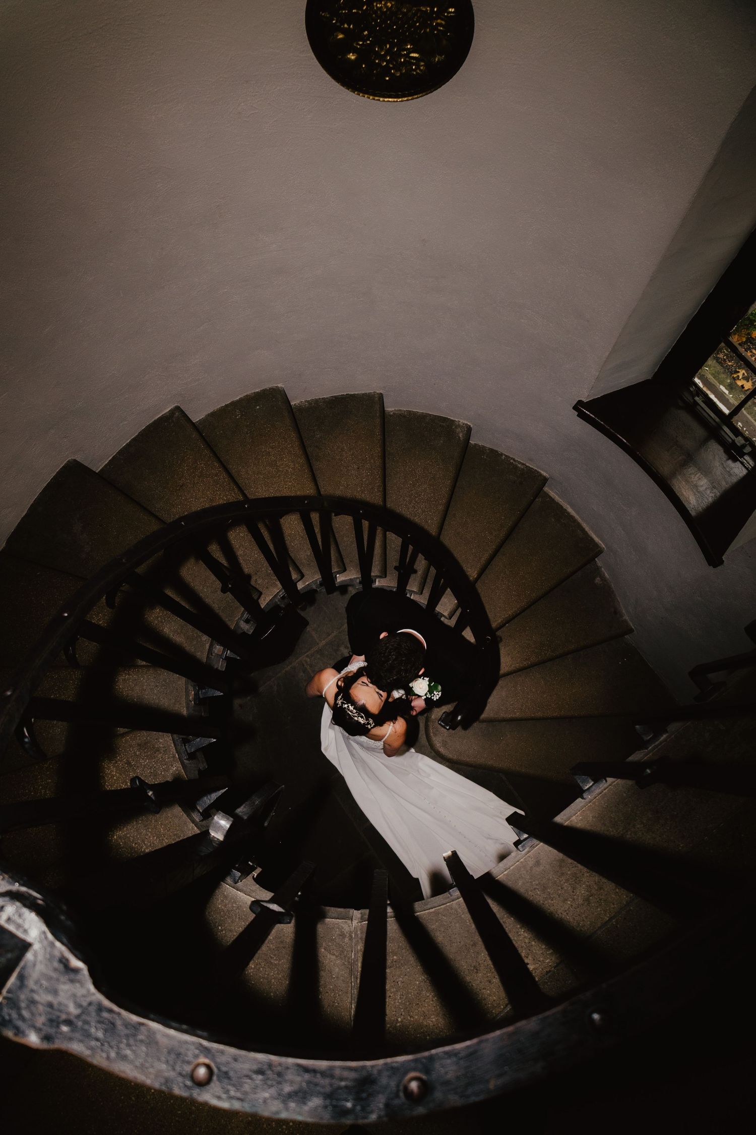 Aerial view of a couple in wedding attire descending a dramatic spiral staircase in a dark interior setting.