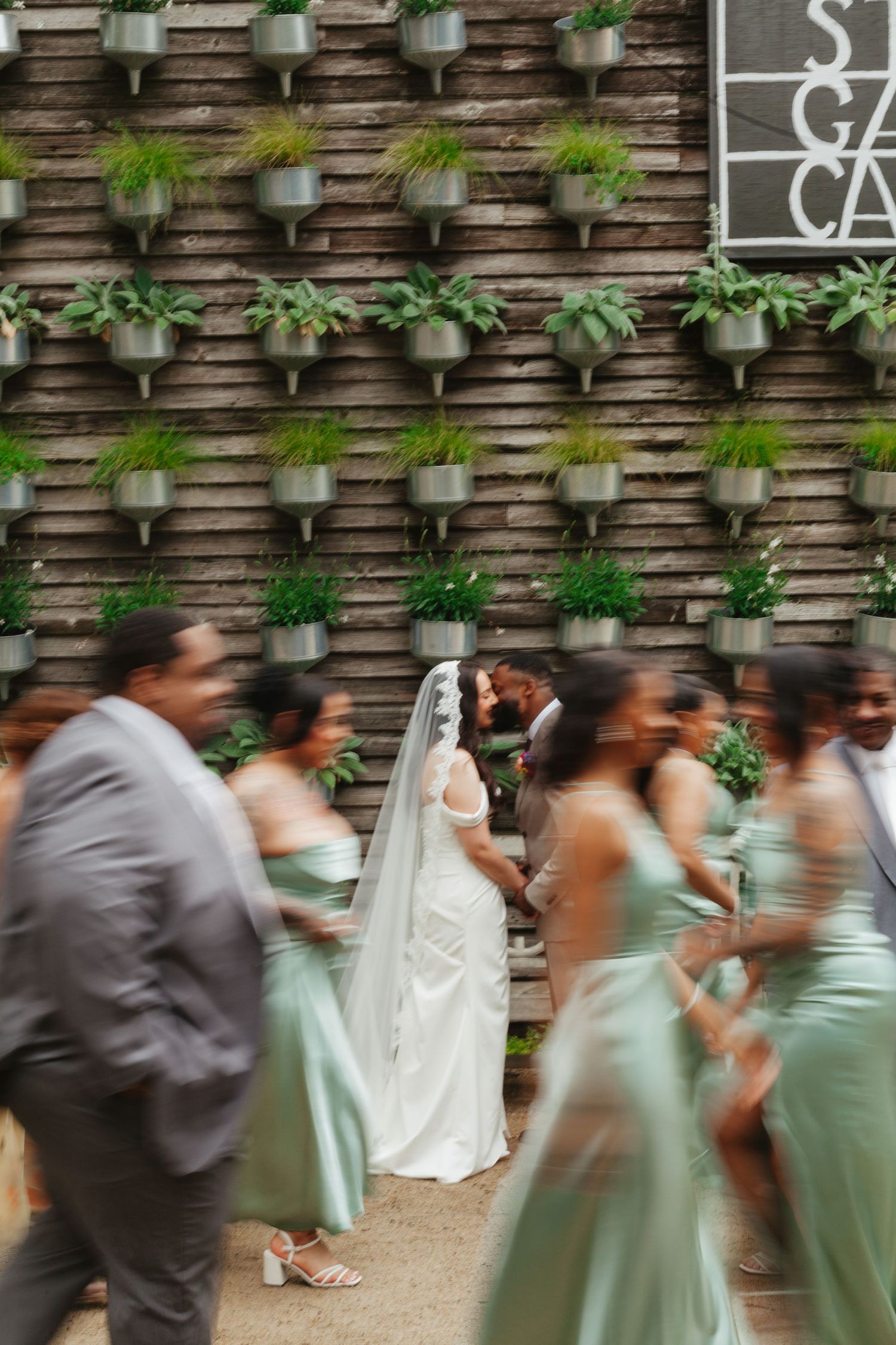 Wedding party members in mint green dresses and gray suits walk in a line against a rustic stone wall with hanging planters.