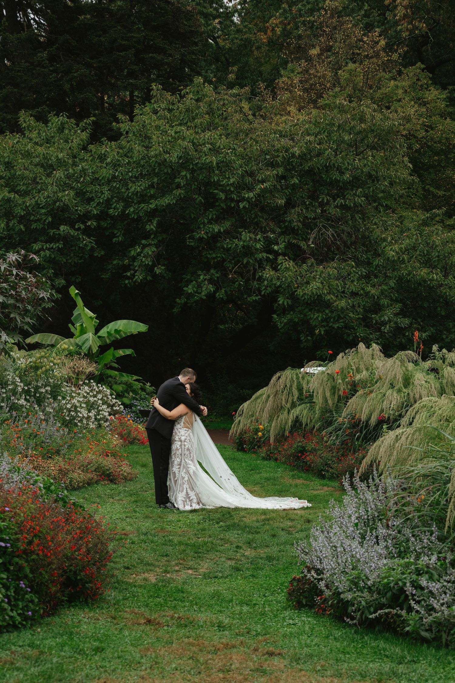 A couple in wedding attire shares romantic moments in a lush garden setting with blooming flowers and greenery.