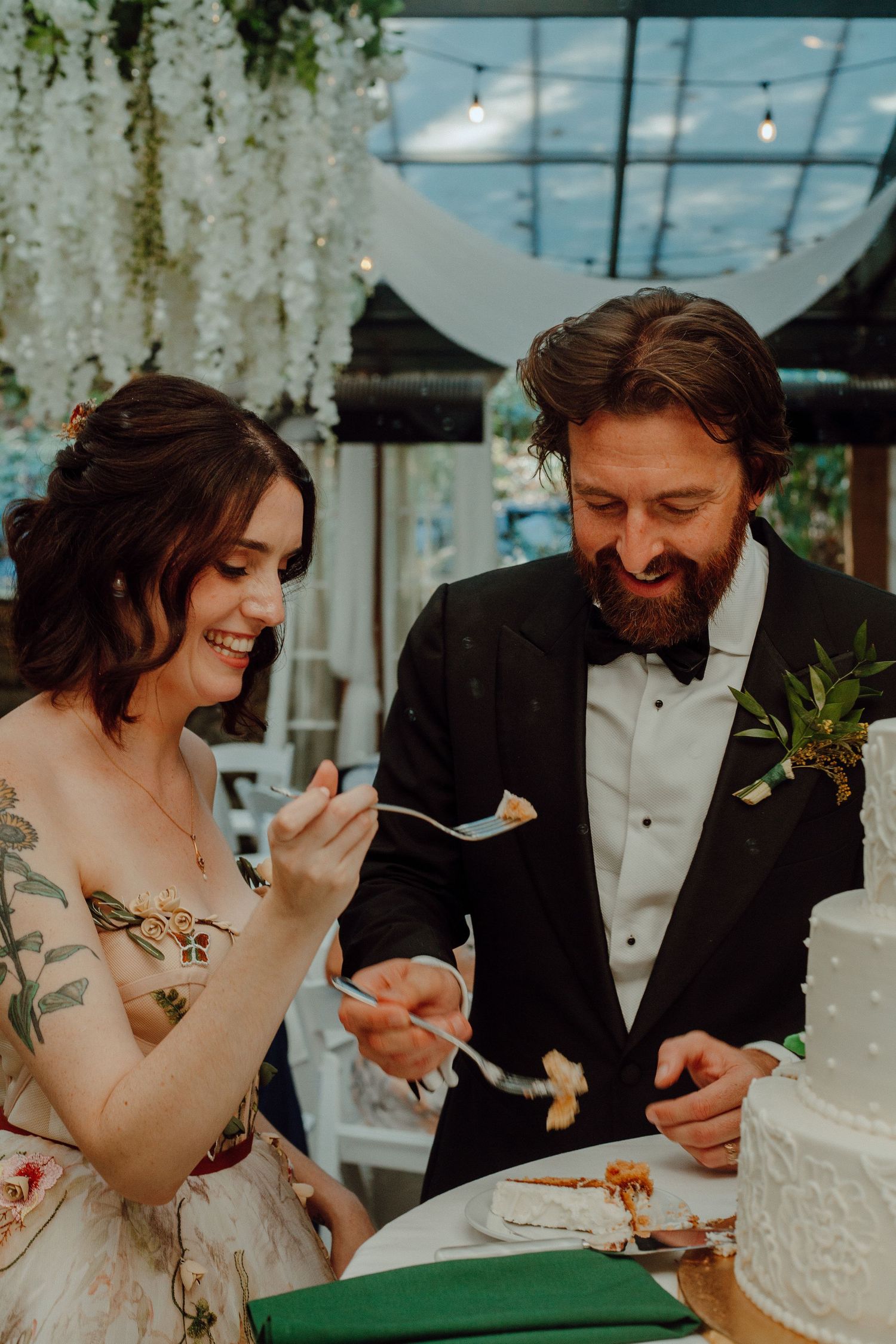 Newlyweds sharing cake at their wedding reception under hanging floral decorations.