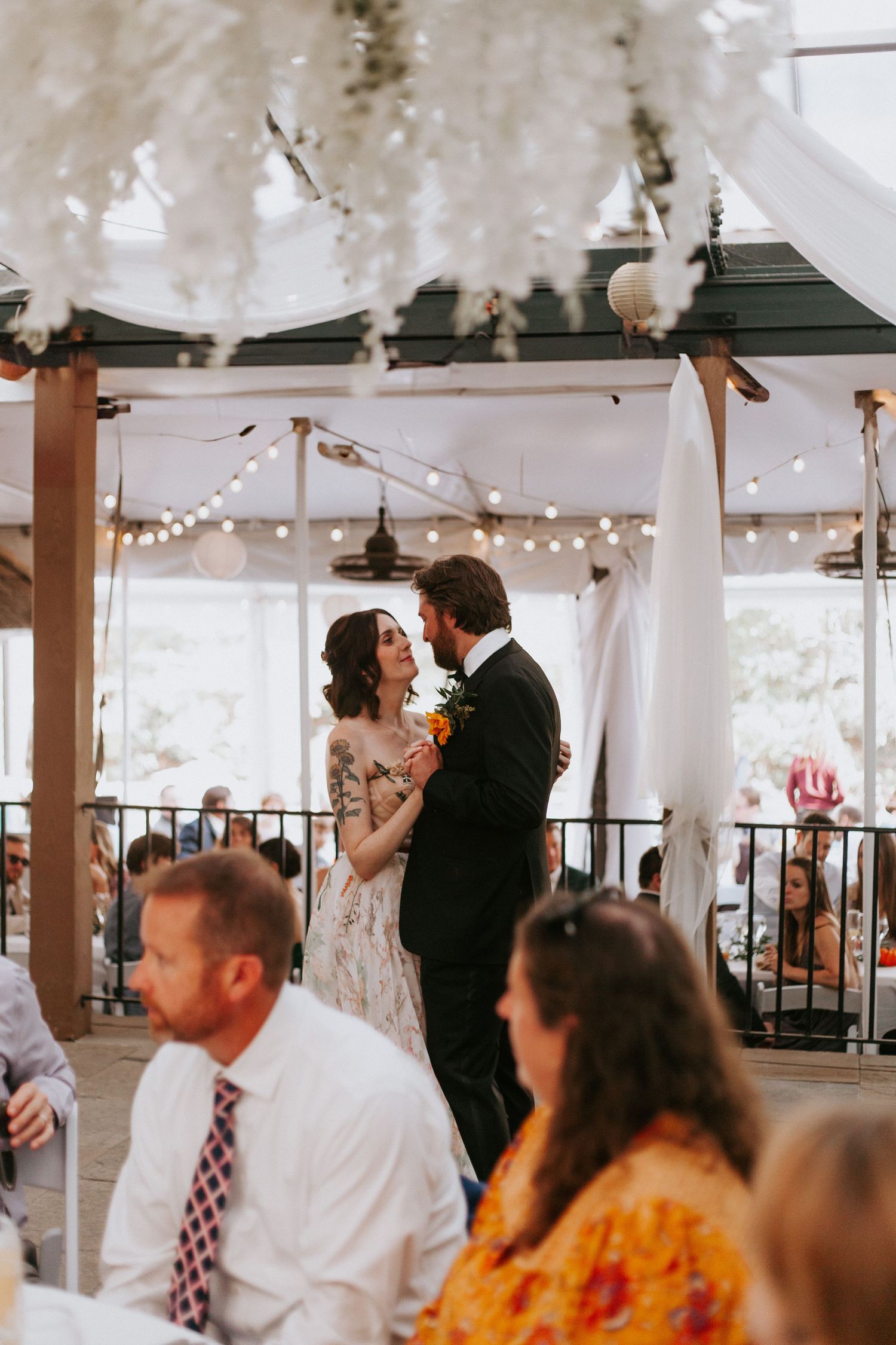 A wedding reception scene with twinkle lights and couples dancing under decorative white fabric.