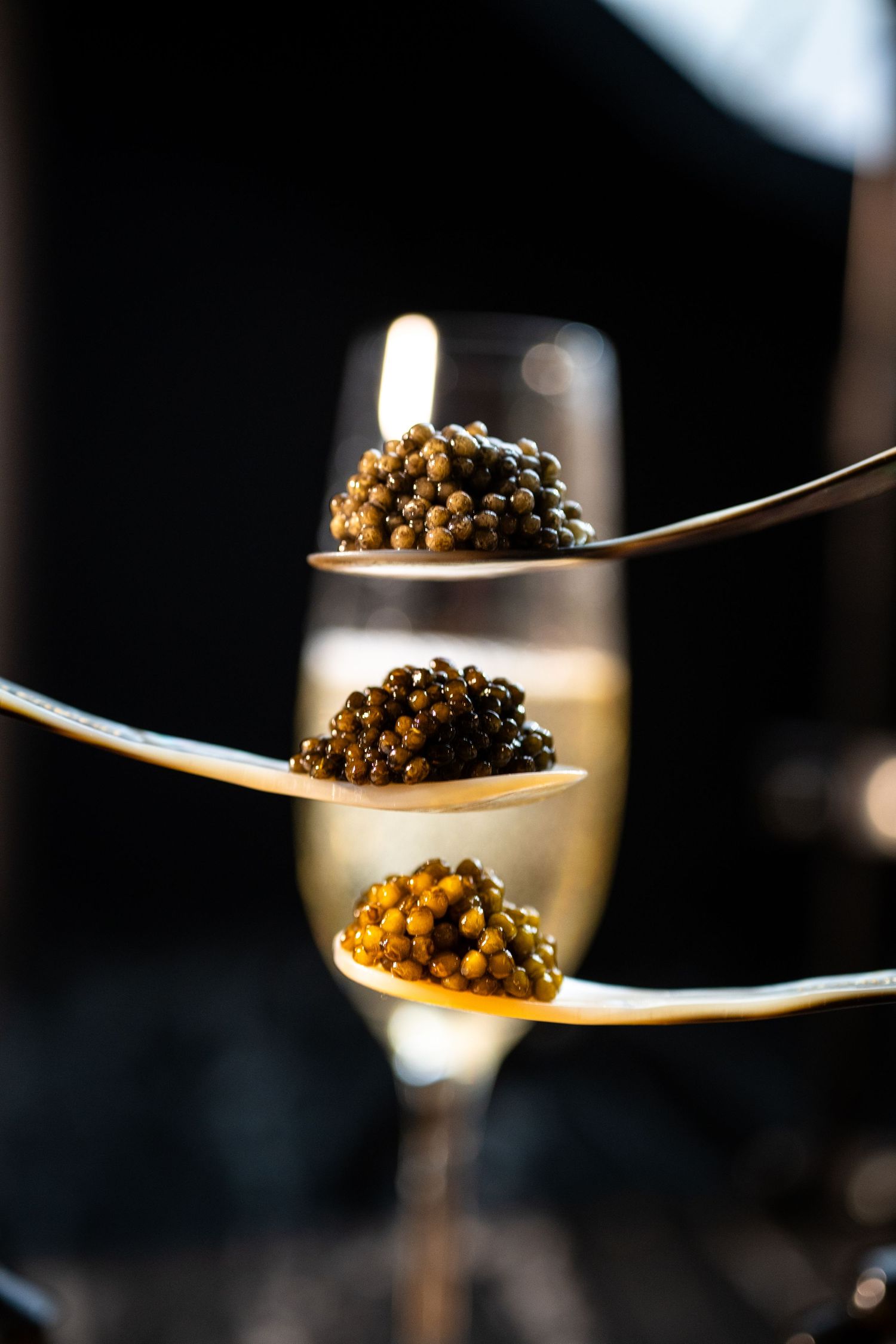 Three golden spoons with black caviar stacked in front of a champagne flute on dark background.