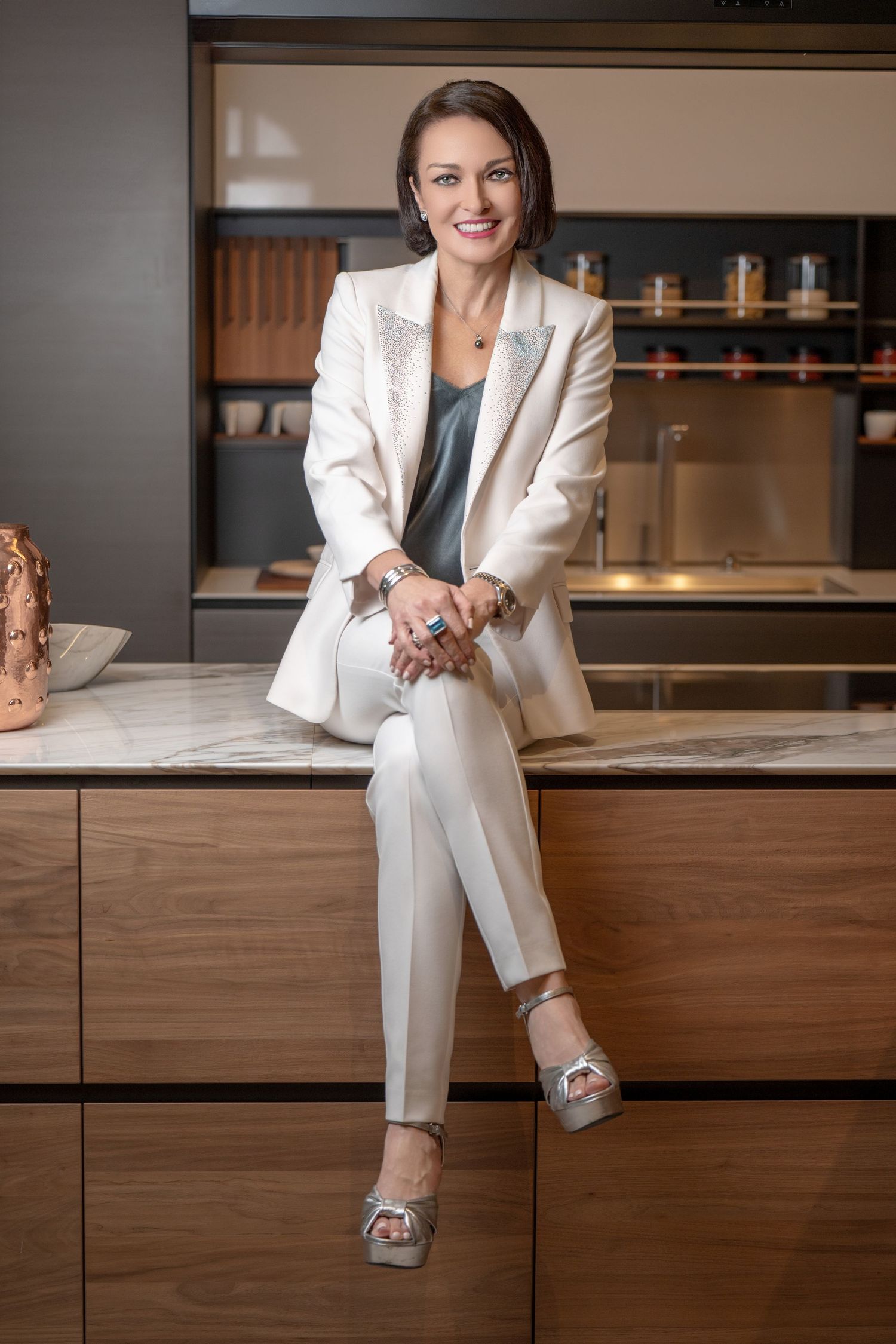 Smiling woman in white blazer and pants sits on marble kitchen counter, wearing silver heels and accessories.