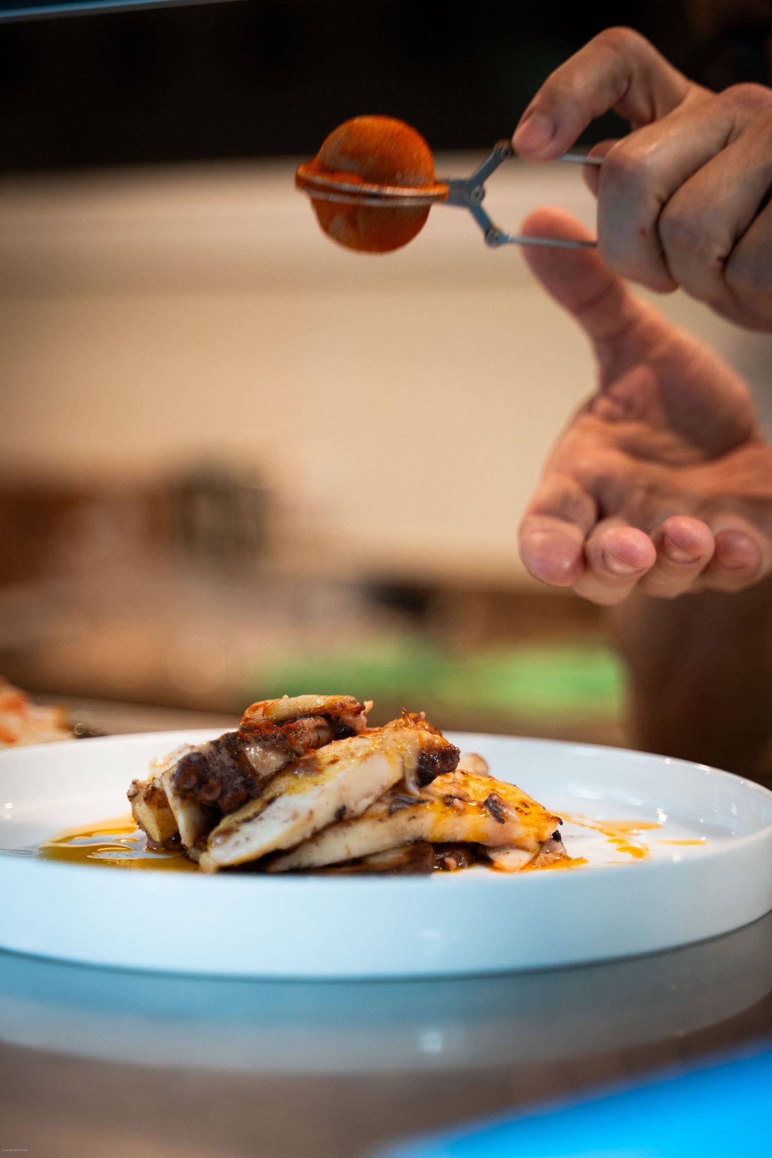 Chef garnishing a plated fish dish with sauce using a spoon in a restaurant kitchen.
