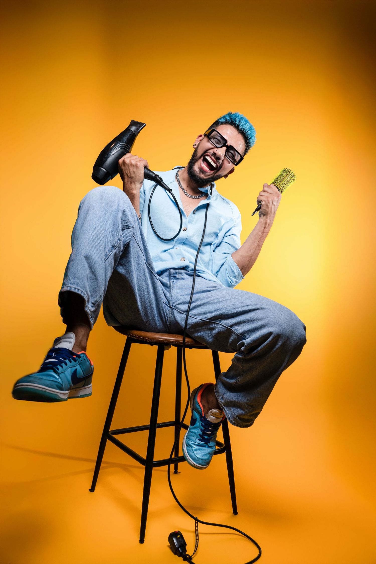 Creative portrait of a professional hairstylist in Houston, Texas, posing with a blow dryer and round brush on a vibrant yellow studio backg