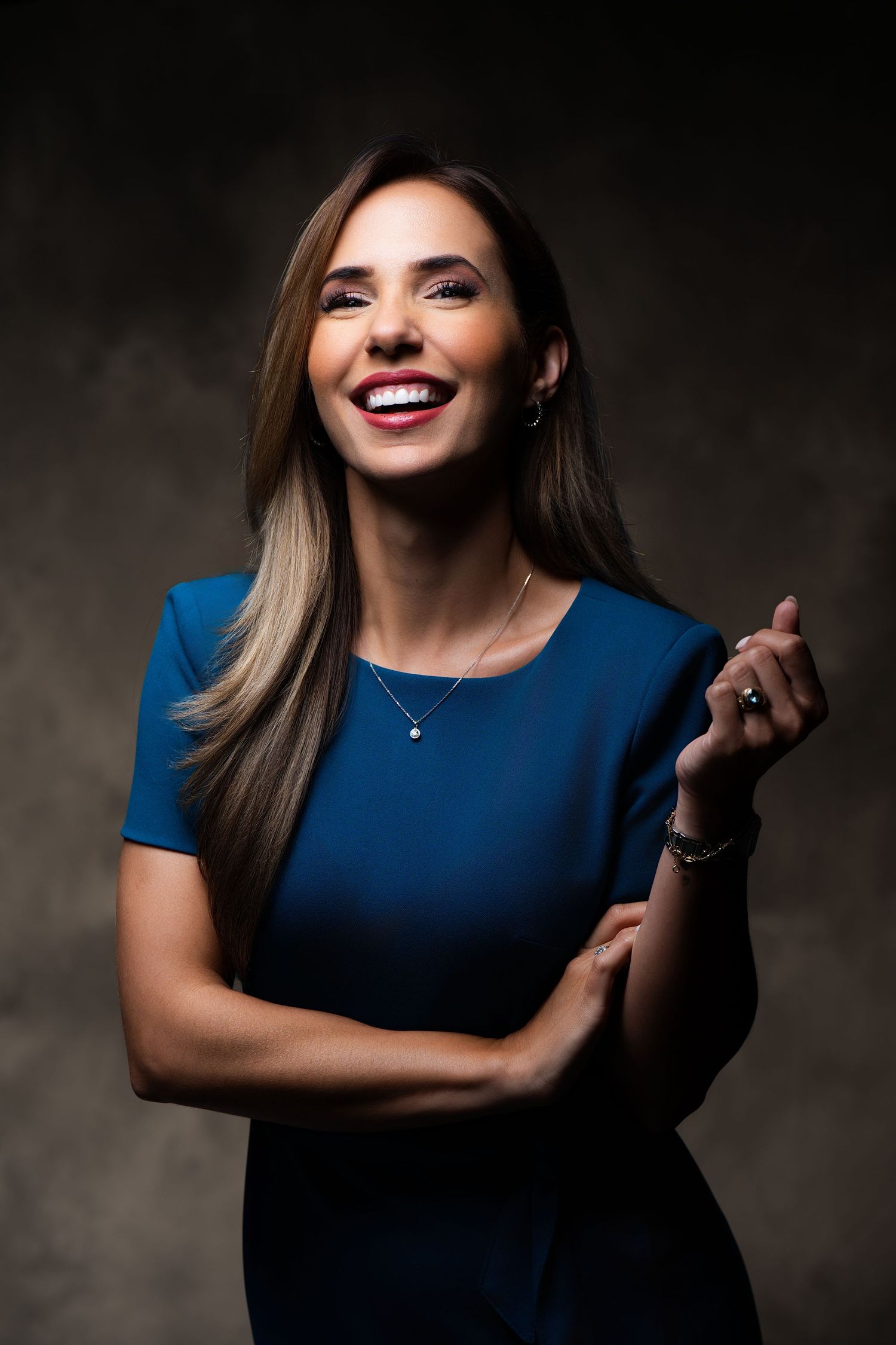 Smiling woman in blue dress with long hair, wearing necklace, posing confidently against dark background.