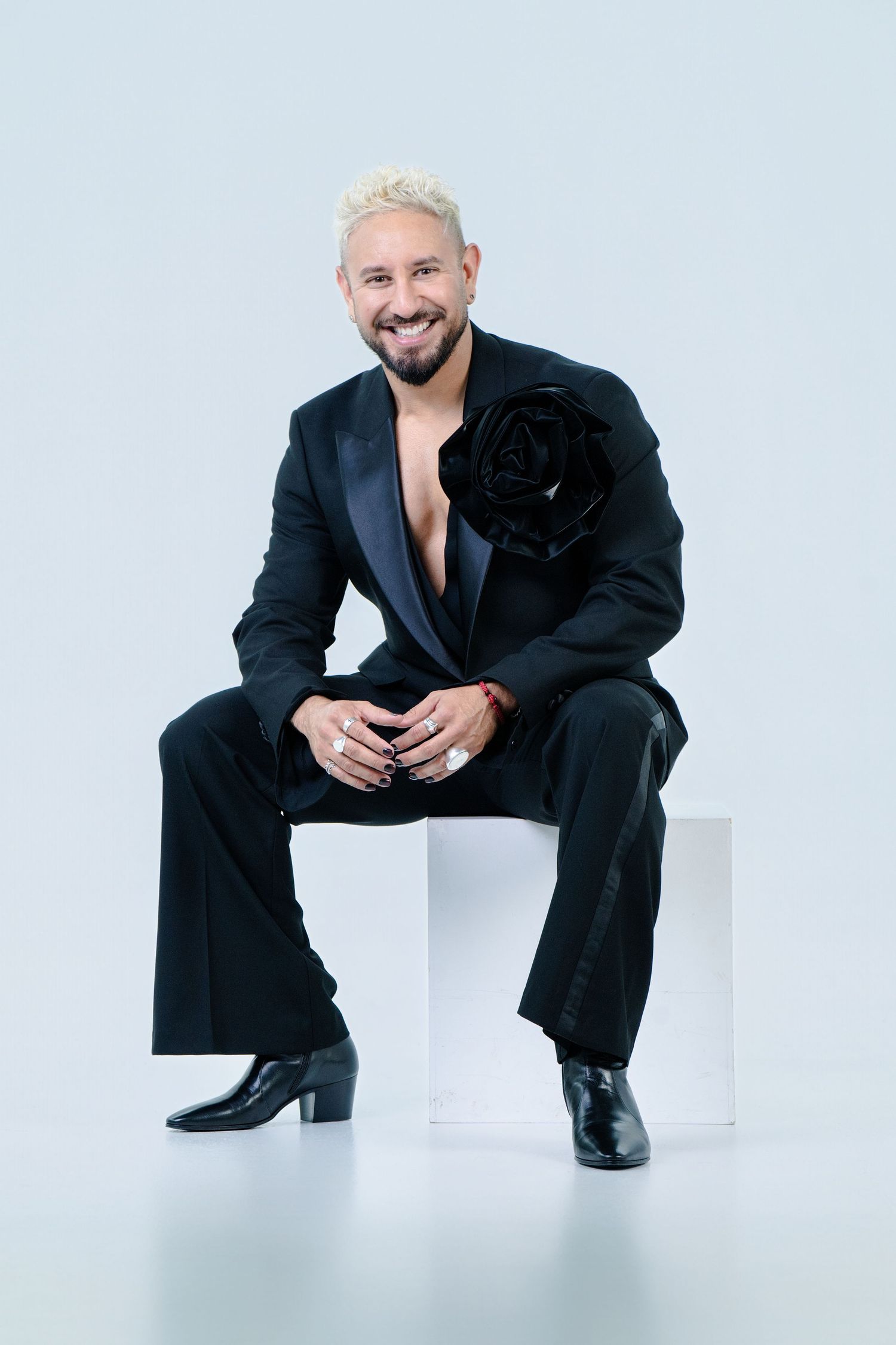 Fashionable figure in black suit and boots sitting on white cube against light backdrop.