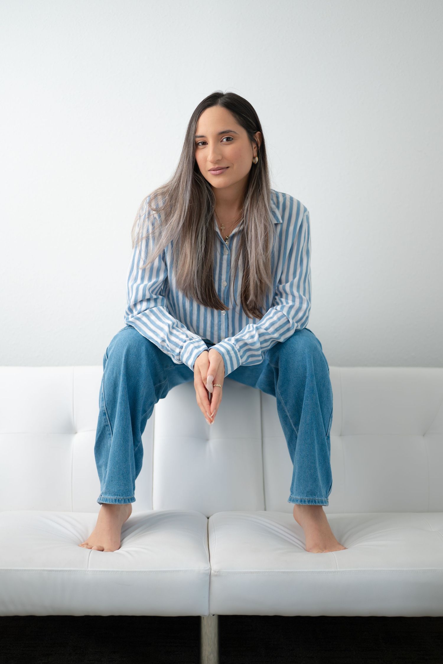Woman in striped shirt and jeans sitting barefoot on white couch against light background.