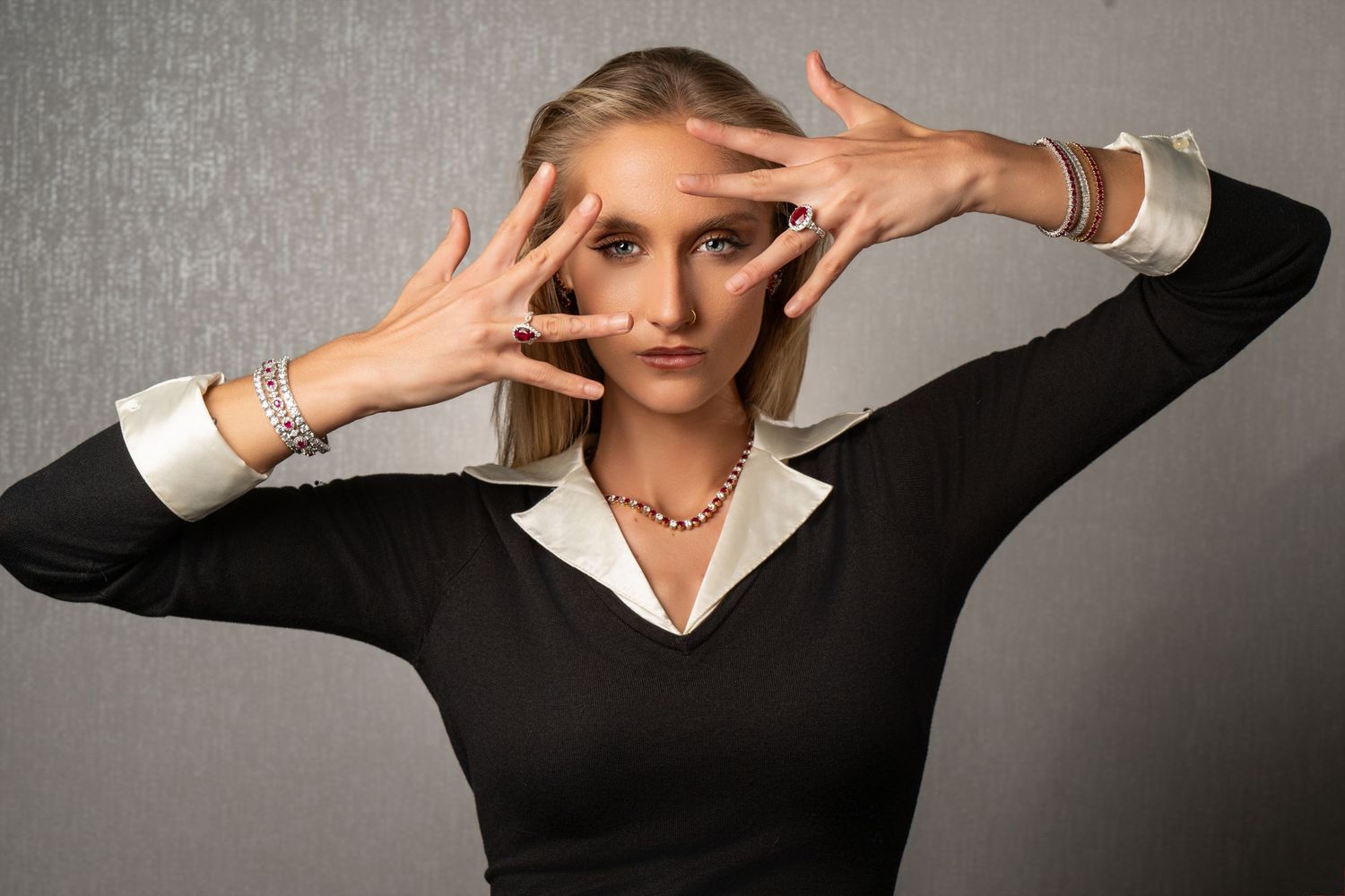 Blonde woman in black top poses with hands framing face, wearing bracelets and pearl necklace.