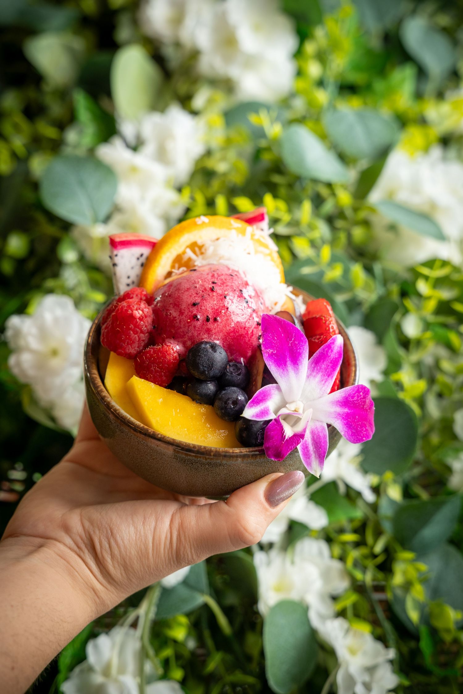 Tropical fruit bowl with dragon fruit, mango and berries garnished with pink orchid against floral background.