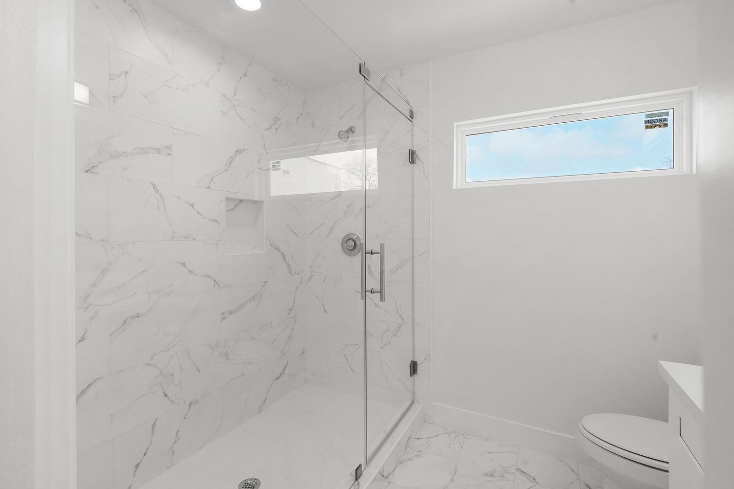 Modern white marble bathroom with glass shower enclosure, chrome fixtures, and natural lighting from window.