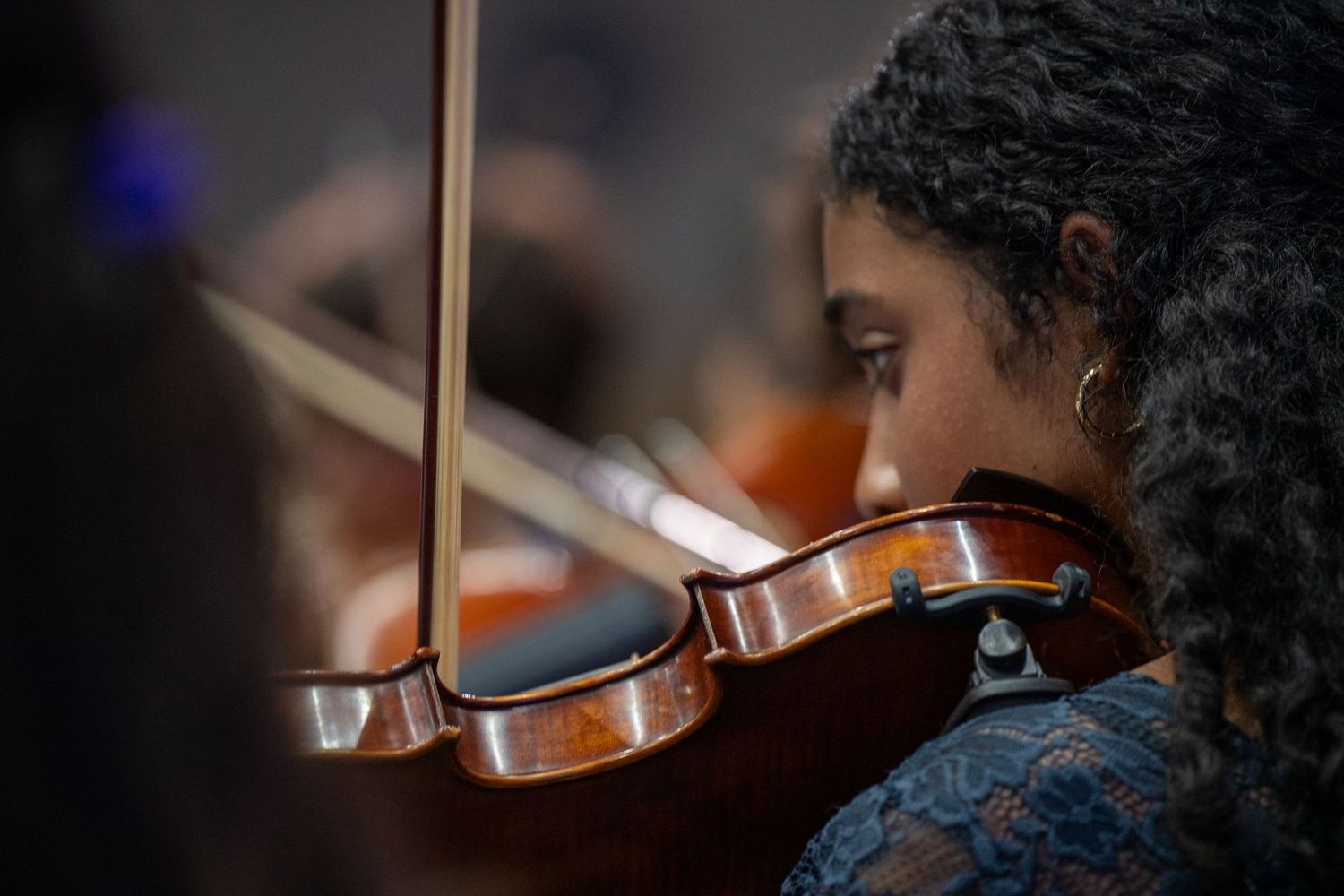 Musician performs with a violin while wearing a blue dress, shown in close-up profile view during a concert.