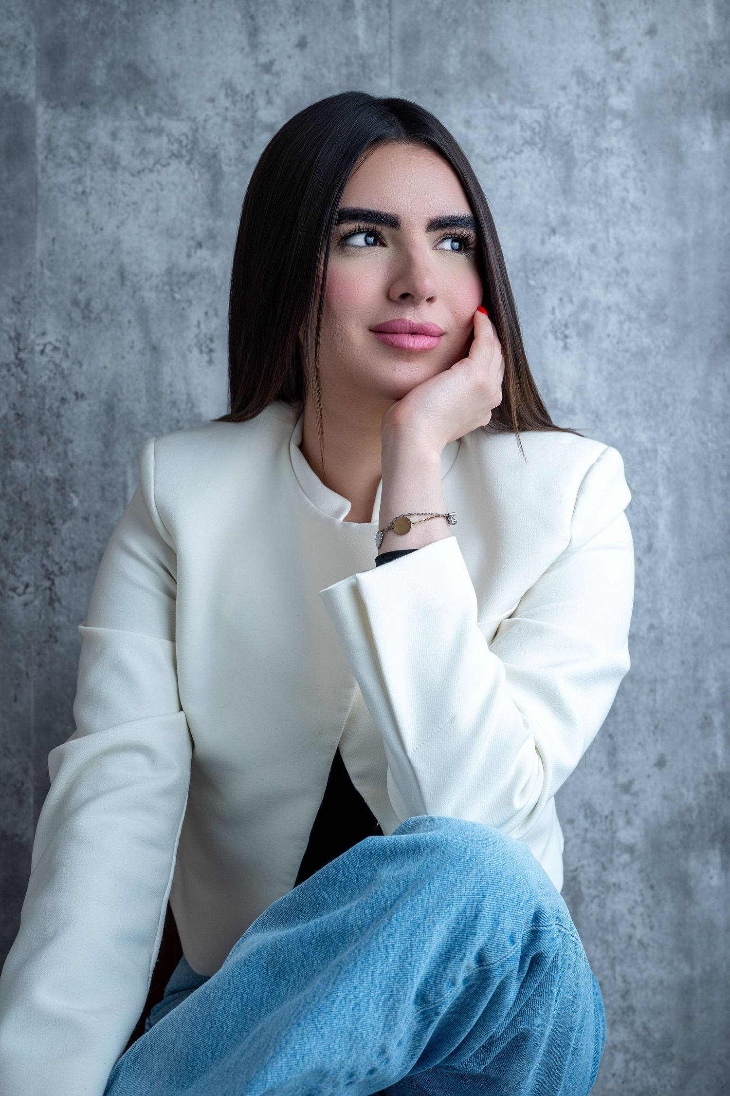 Fashion portrait of a person in a white blazer and blue jeans against a gray concrete wall background.