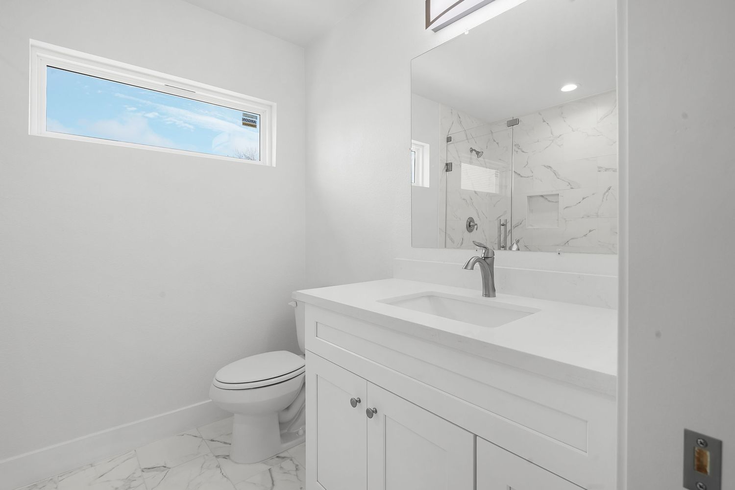 A modern white bathroom interior featuring marble tile, floating vanity, and natural light from transom windows.