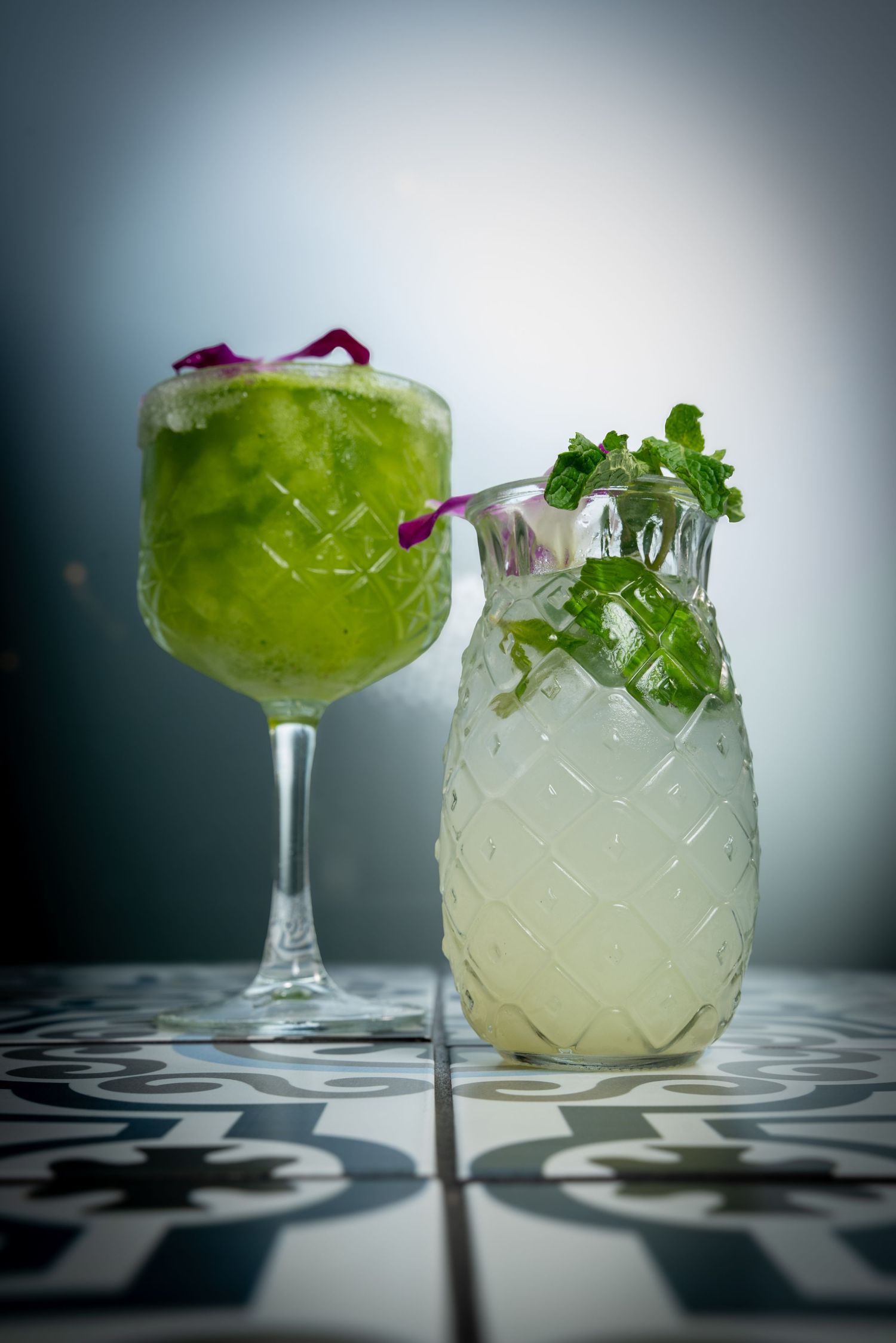 Two elegant cocktails garnished with herbs and flowers on a patterned surface.