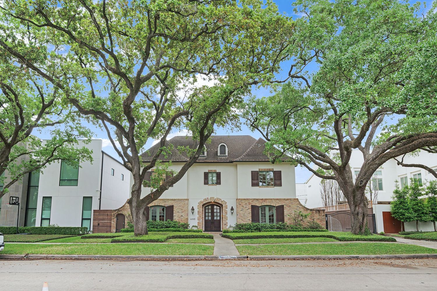 White two-story colonial home with large mature oak trees lining the front yard on a sunny day.