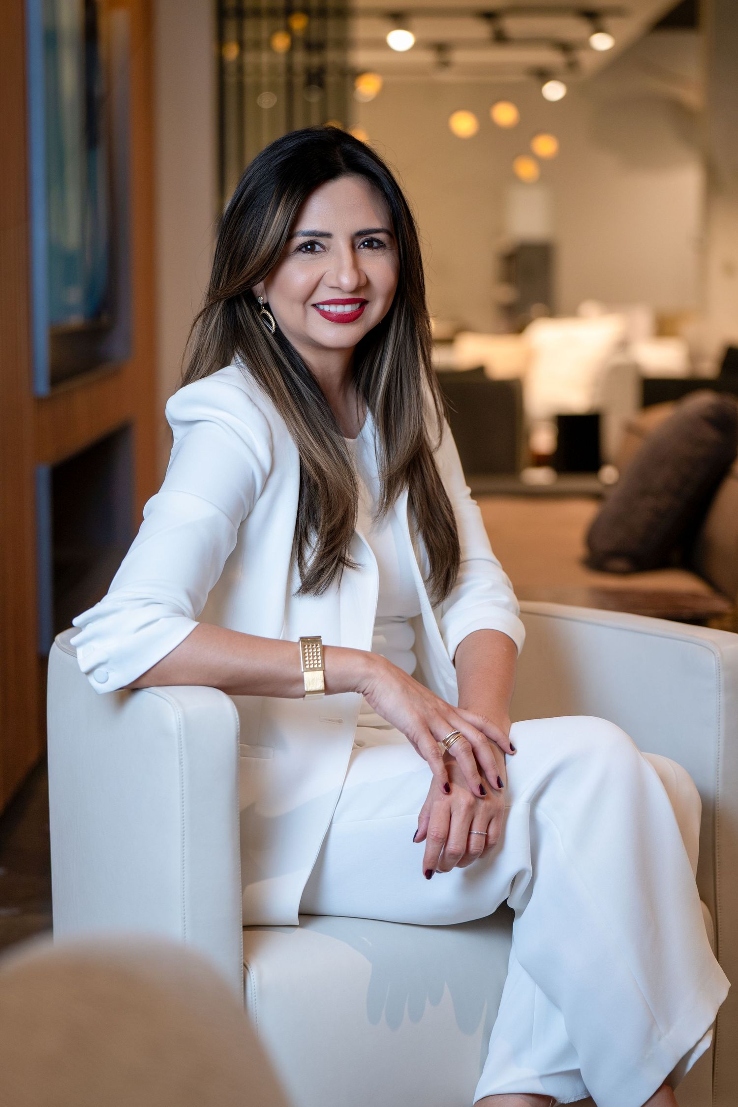 Professional wearing all white business attire sits elegantly on white chair in modern office setting with warm lighting.