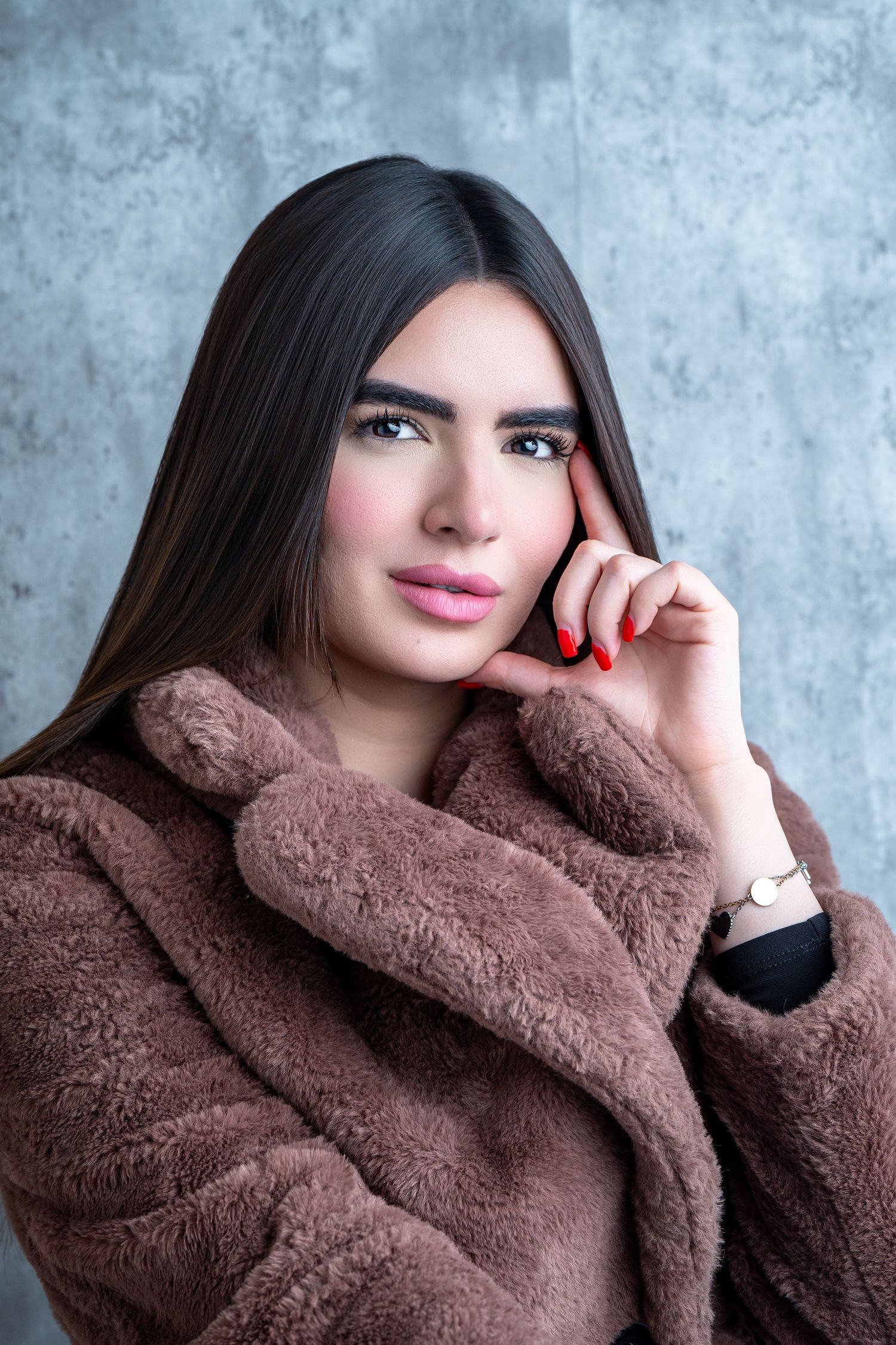 A person wearing a cozy brown faux fur coat poses against a gray textured background in a stylish portrait series.