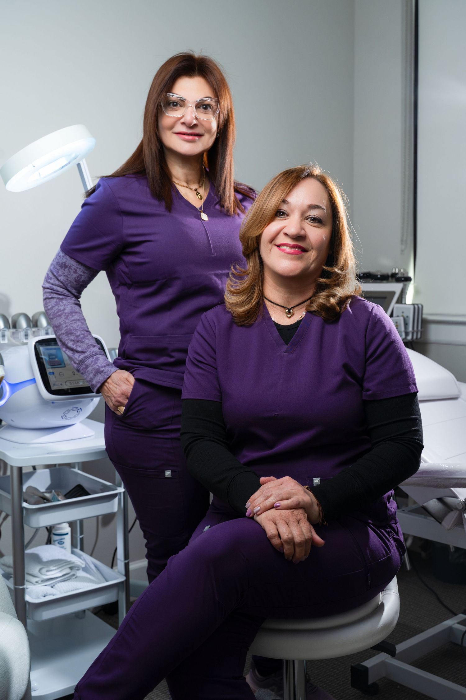 Two female medical professionals in purple scrubs pose together in a modern clinical treatment room with medical equipment.