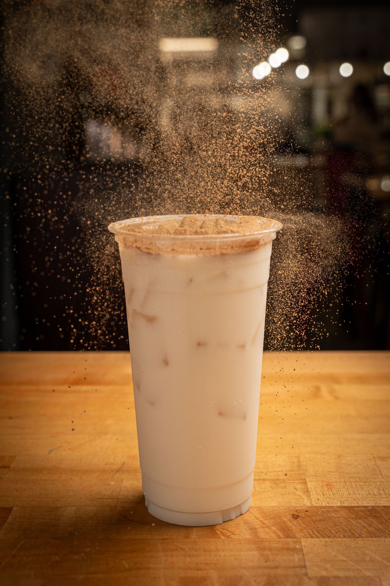 Iced milk drink in a large plastic cup with cinnamon powder being sprinkled on top, on a wooden table.
