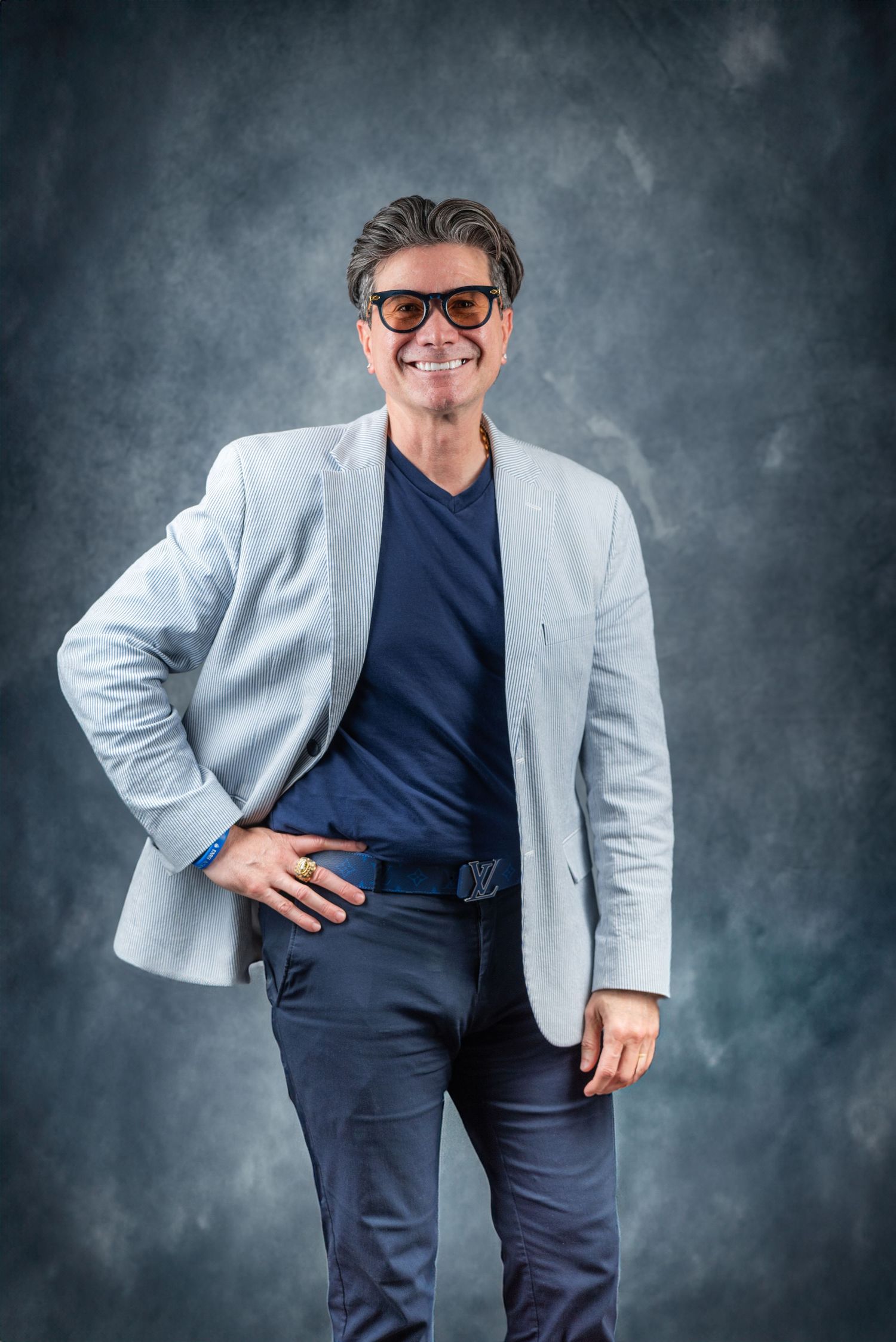 Smiling middle-aged man in light gray blazer, navy shirt, and sunglasses posing confidently against a dark studio background.
