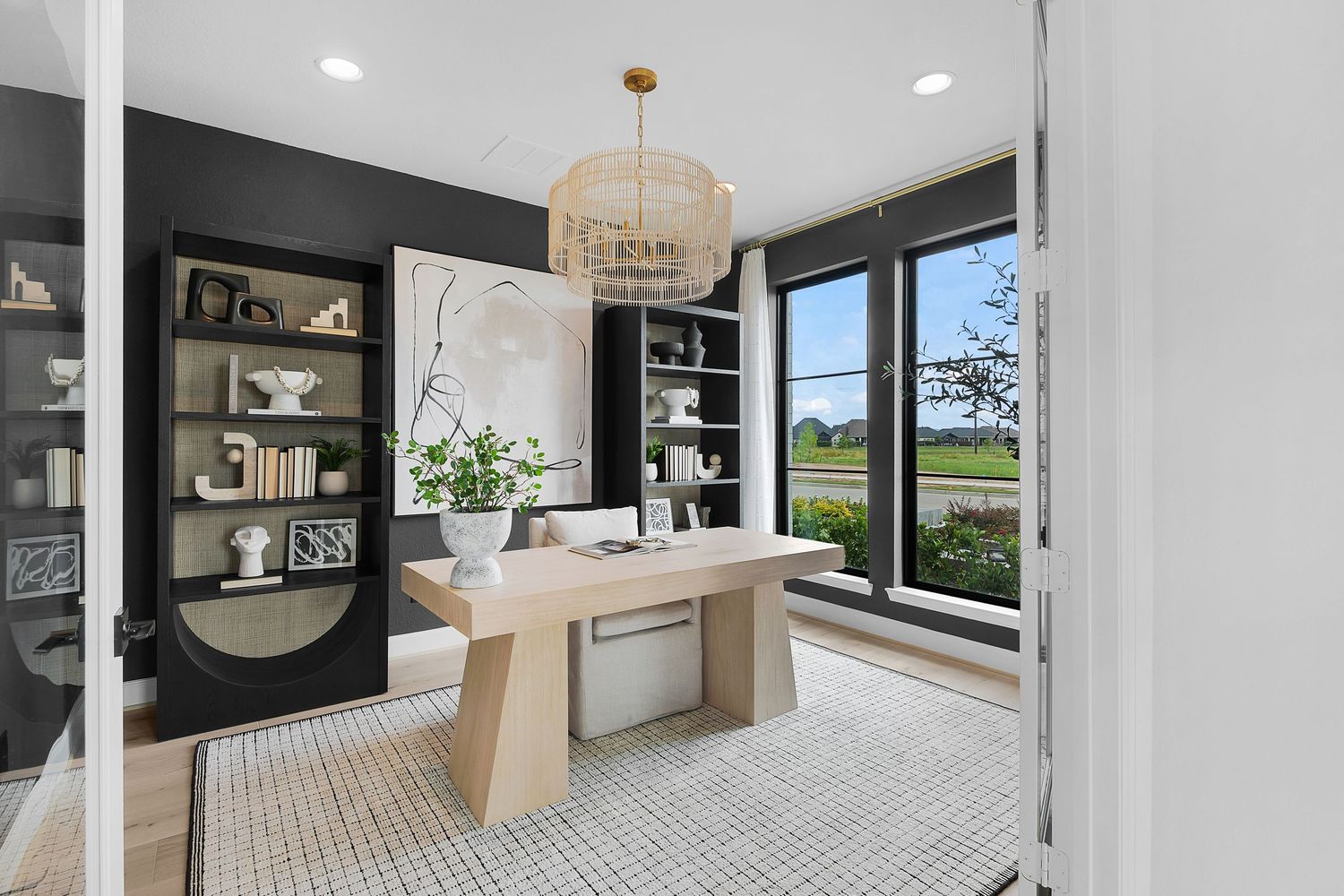 Modern home office featuring a light wood desk, black built-in shelving, and a contemporary beaded chandelier.