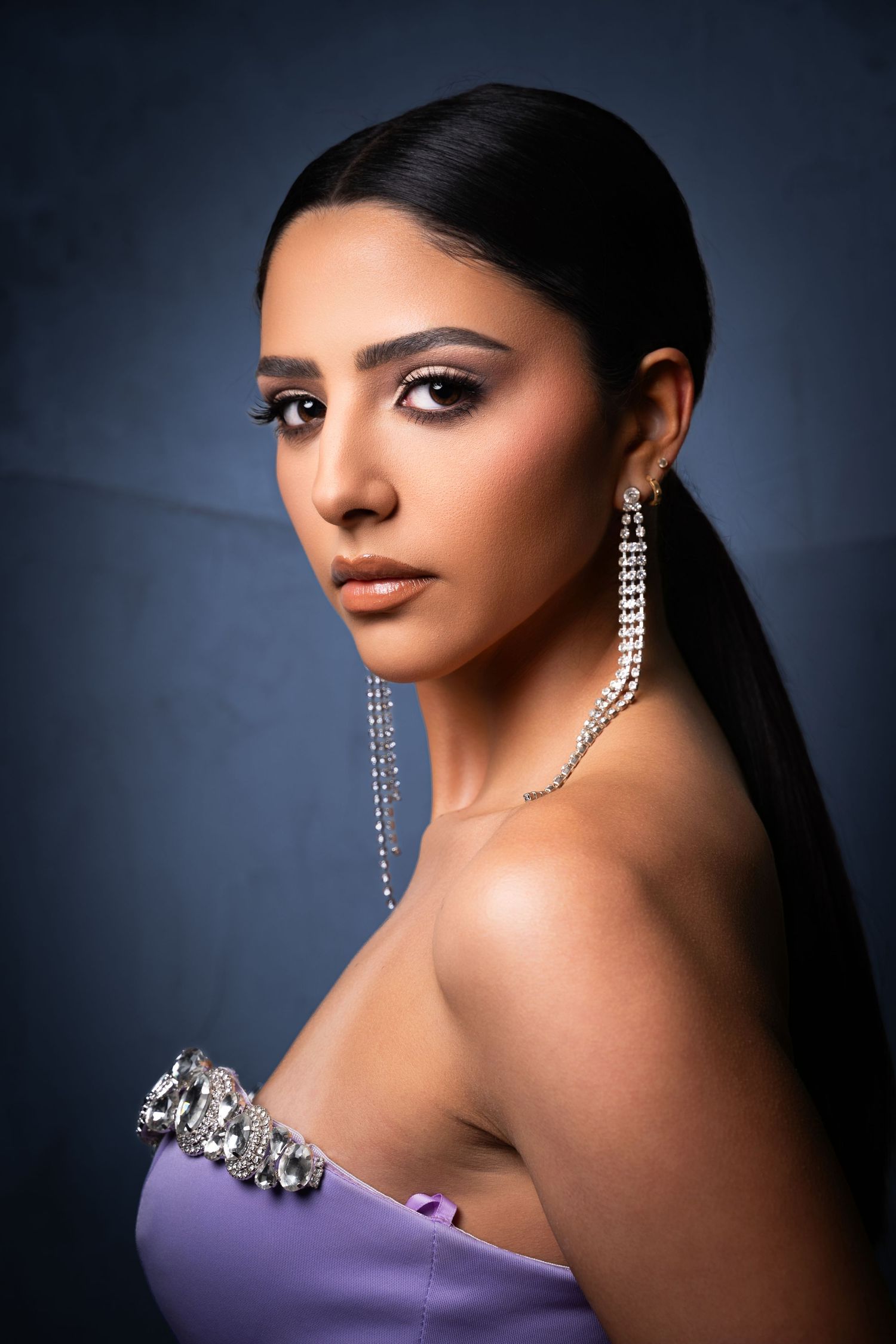 Portrait against dark background in purple gown with long diamond earrings and sleek black hair.