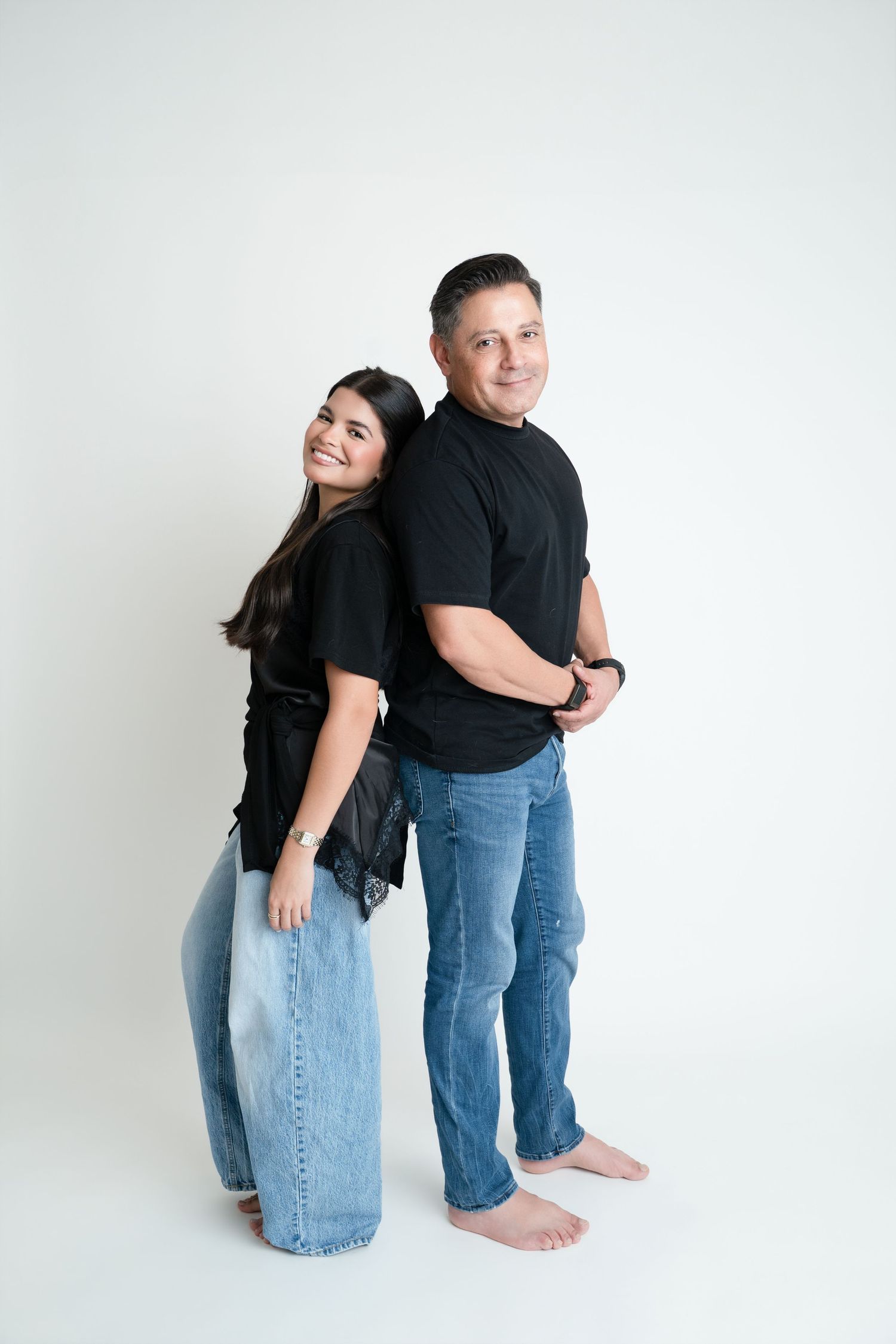 A man and woman in matching black shirts and jeans pose back-to-back smiling in a white studio.