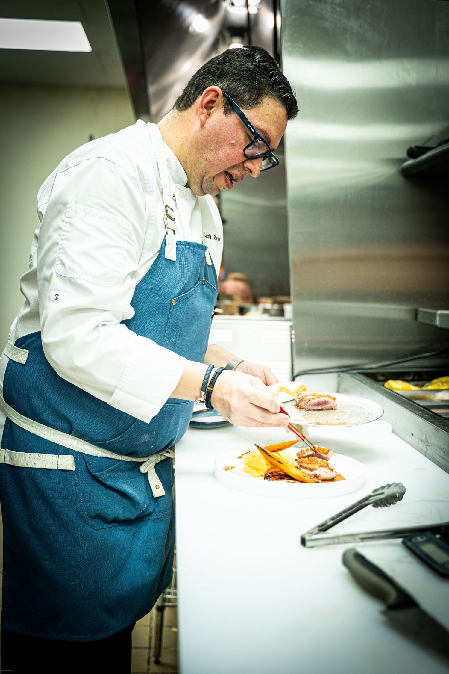 Chef in white coat and blue apron preparing food in a professional kitchen.