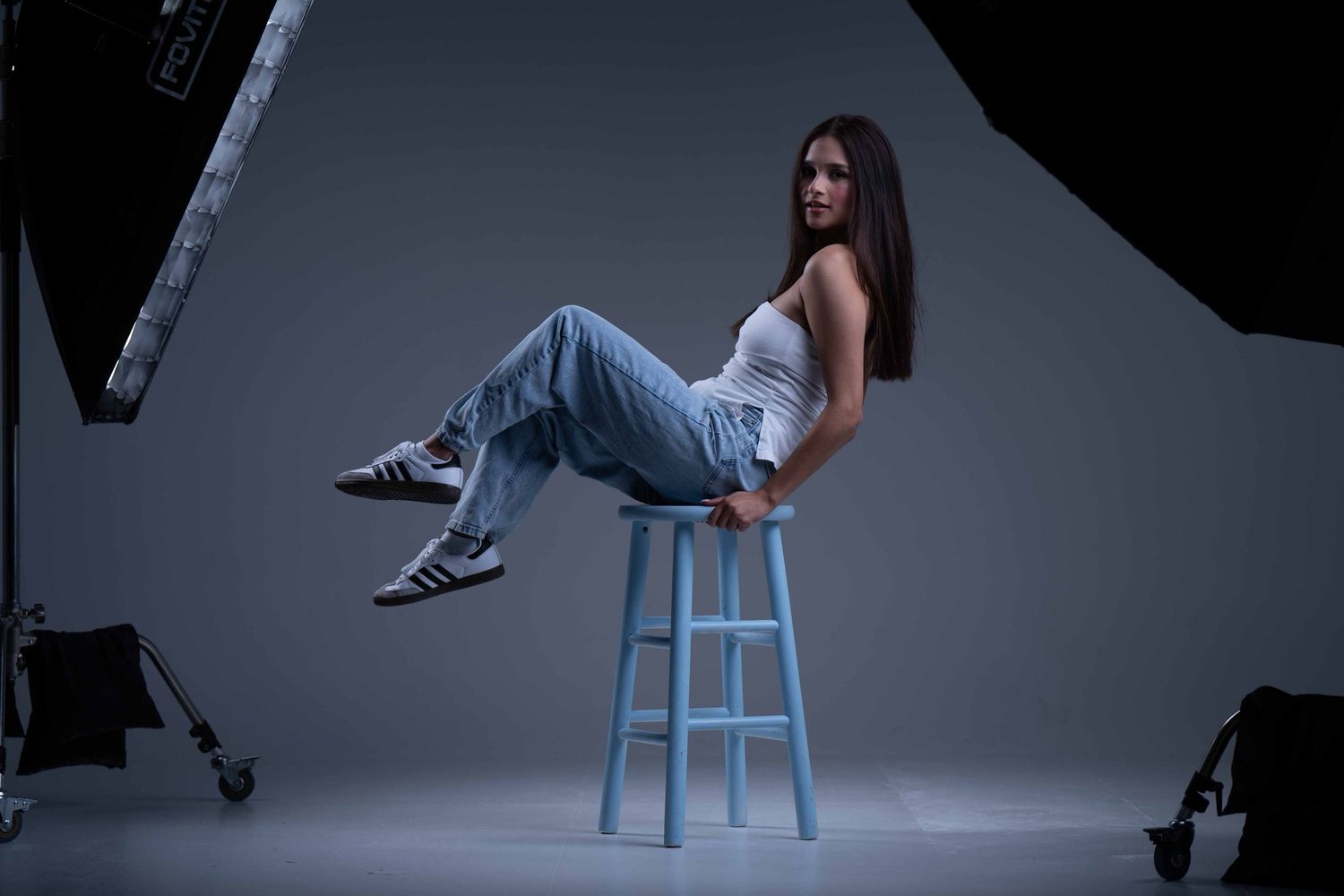 Young woman in jeans and white top posing on a blue stool in a professional photography studio.