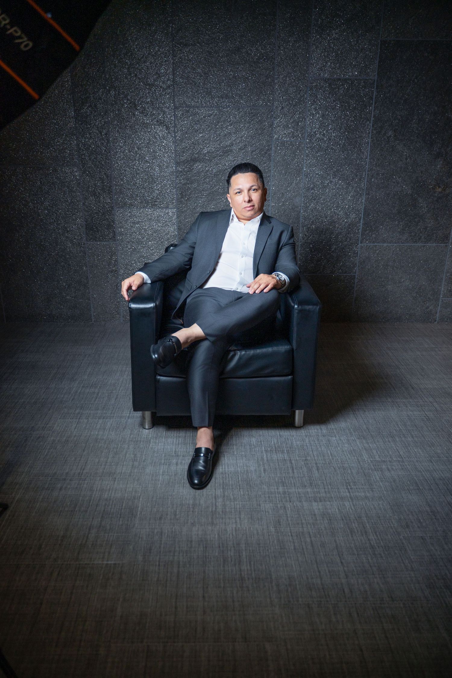 Professional figure in dark suit reclining on black leather chair against textured gray wall in moody lighting.