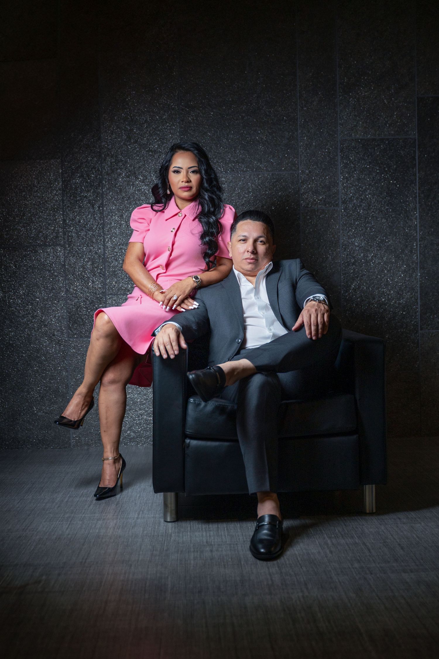 Dark dramatic photo series showing two people in contrasting outfits - pink dress and gray suit - posed on black leather couch.