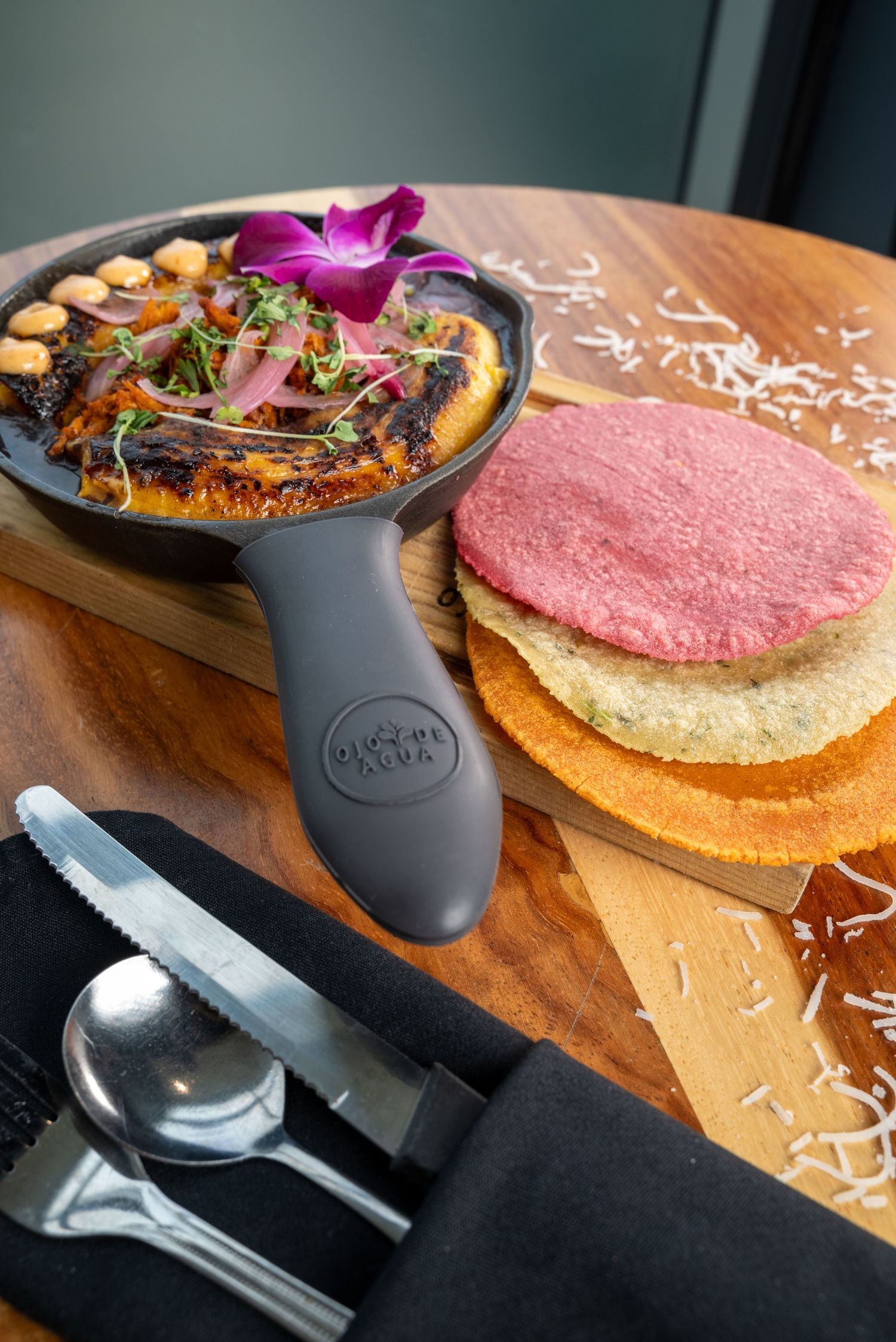 Cast iron pan with garnished dish and colorful tortillas on wooden serving board.