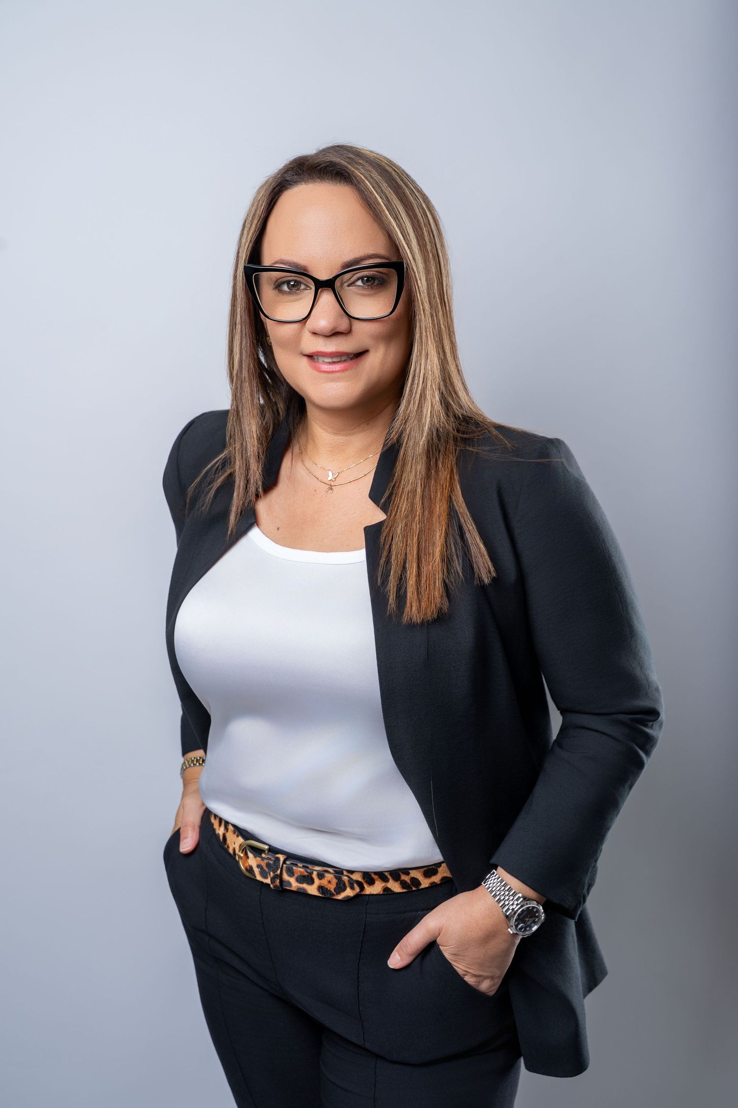Professional headshot of someone wearing a black blazer, white top, and glasses against a gray studio backdrop.