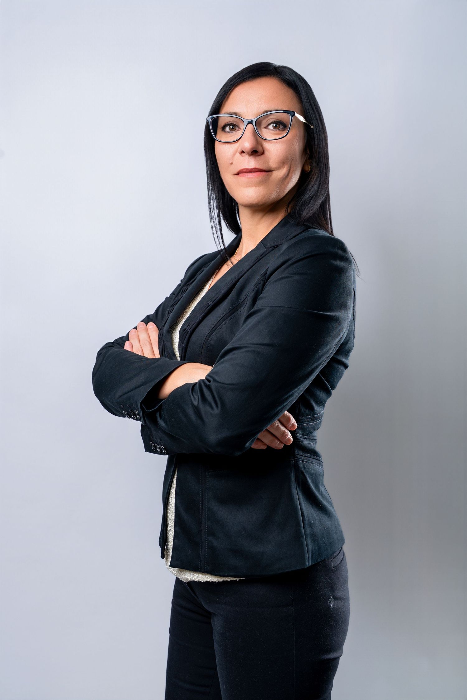 Professional in a black business suit and glasses stands confidently with arms crossed against gray background.
