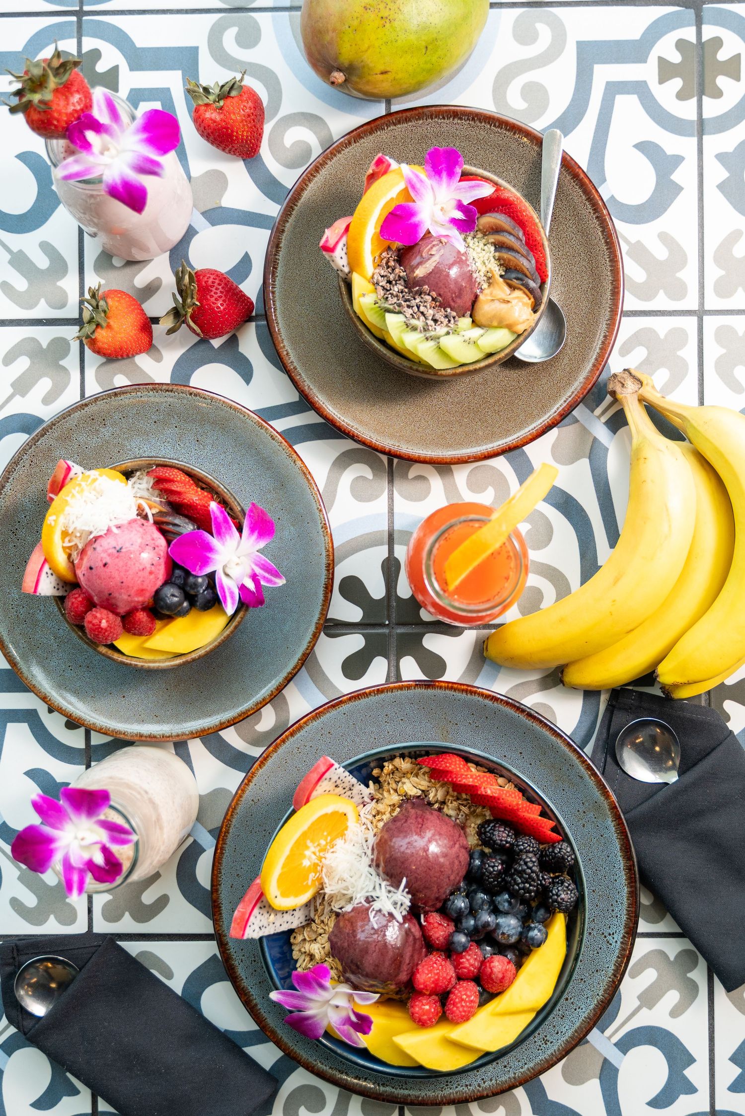 Overhead view of acai bowls with fresh fruits and orchid flowers on patterned tile table.