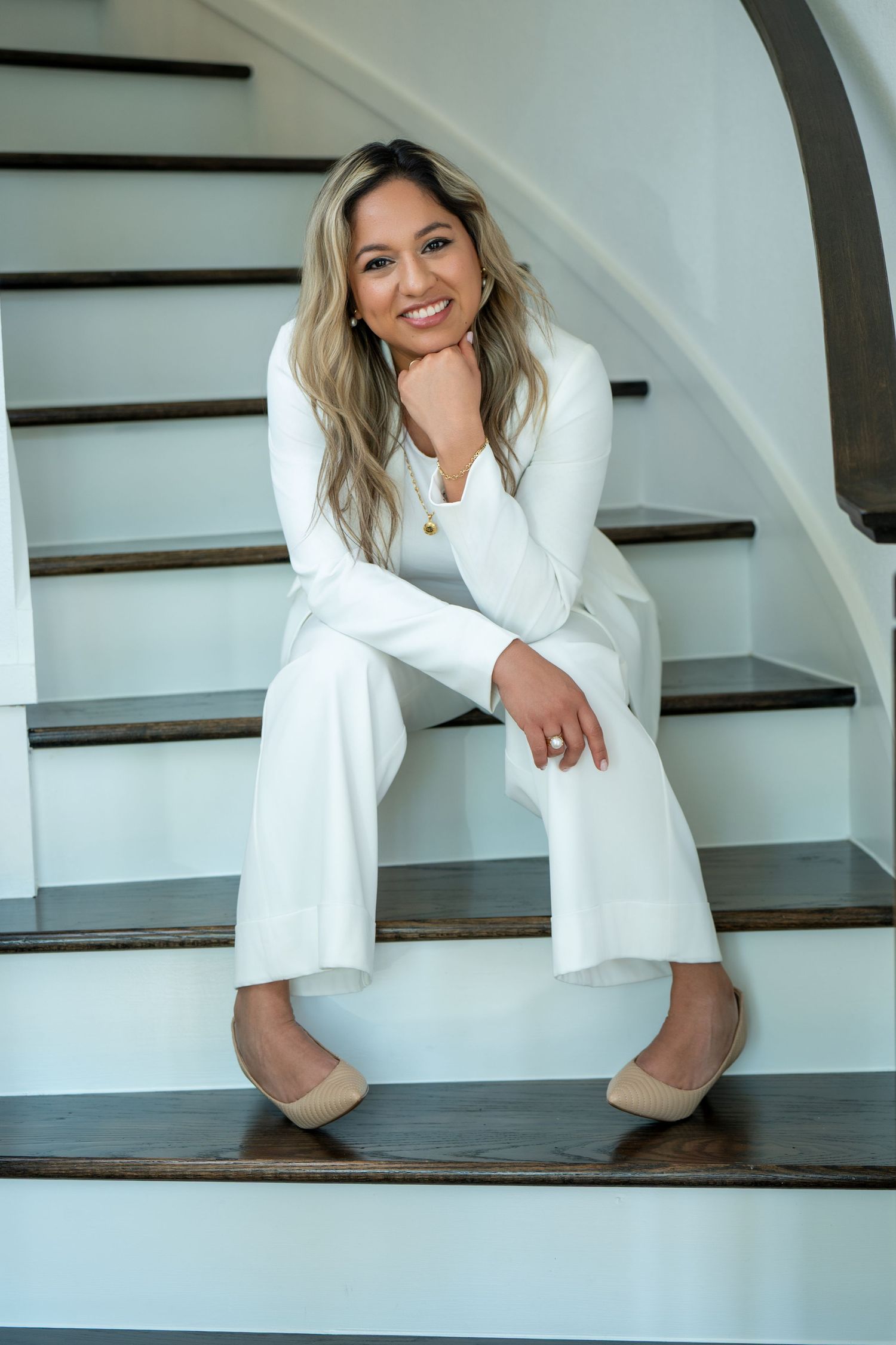 Person in white blazer sits casually on dark wooden stairs in bright interior setting.