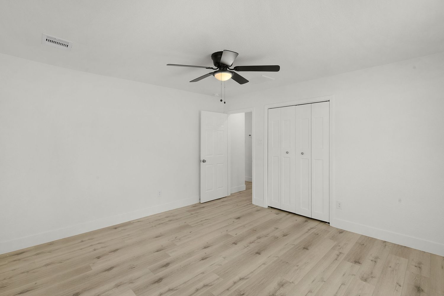 Empty bedroom with white walls, ceiling fan, and light wood flooring showing clean minimalist design.