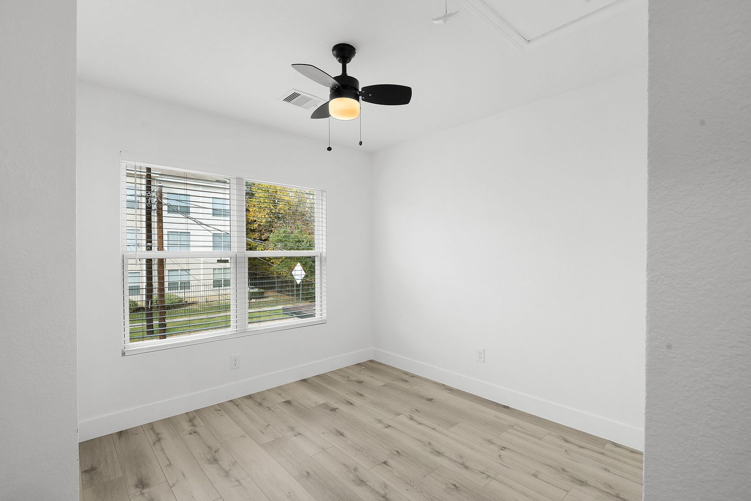 Modern room interior with light wood flooring, white walls, ceiling fan, and large windows providing natural light.