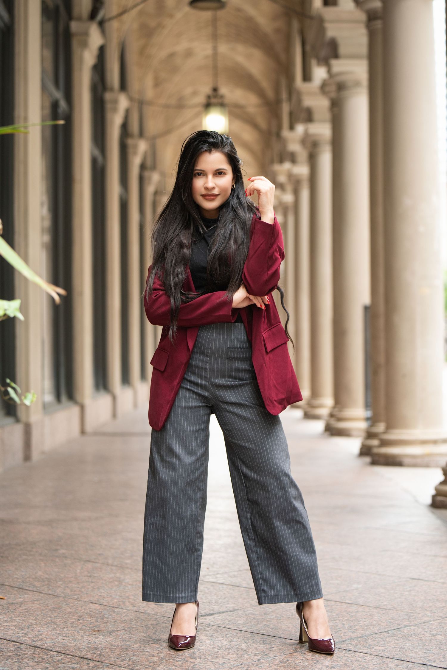 Person in burgundy blazer and striped pants posing in elegant colonnade.