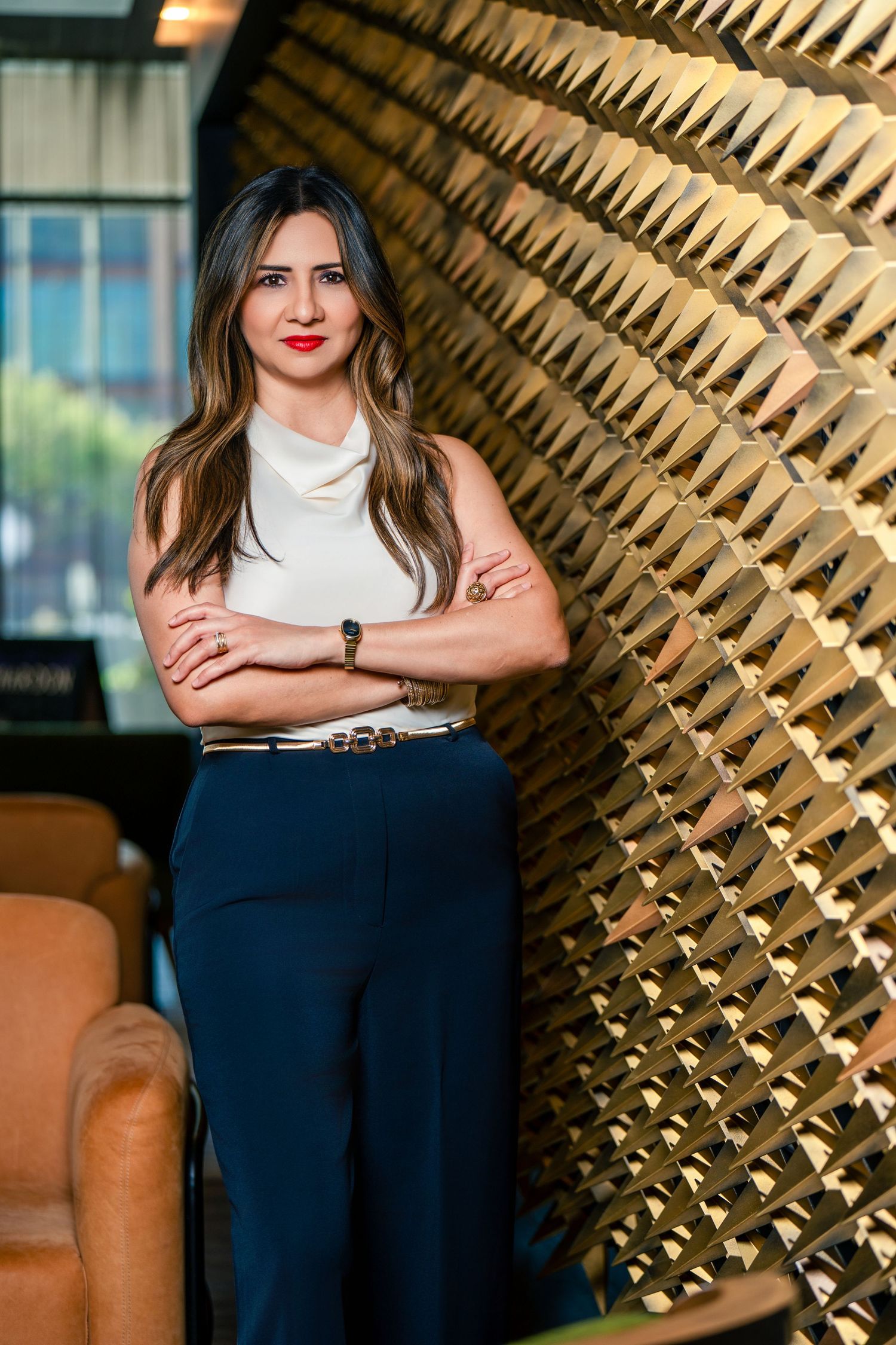 Professional woman in white top and navy pants stands confidently with arms crossed against a gold geometric wall.